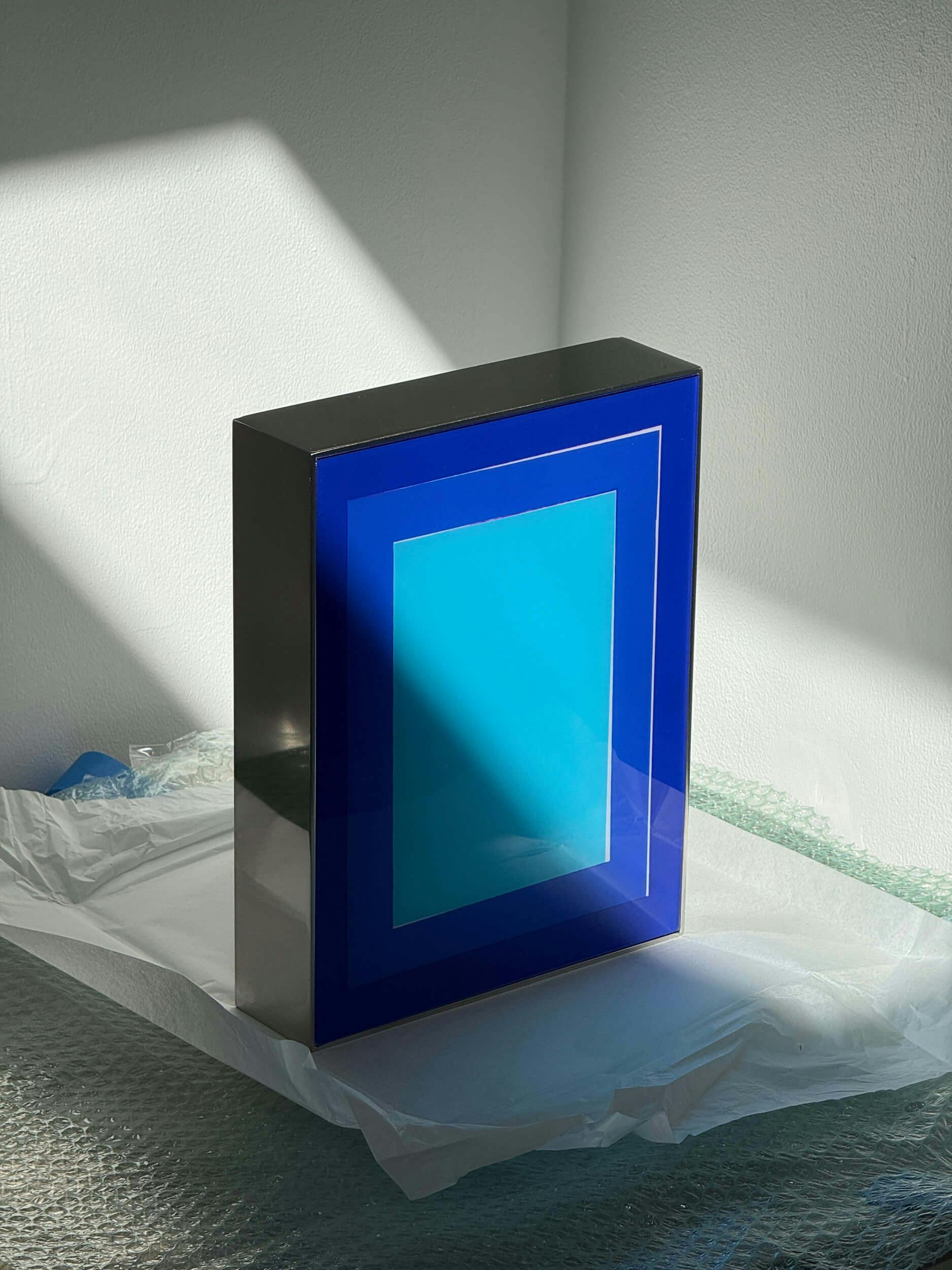 A rectangular object with a glossy surface featuring a geometric design in blue and light blue stands on a covered surface. Sunlight casts angular shadows on the white wall and object.
