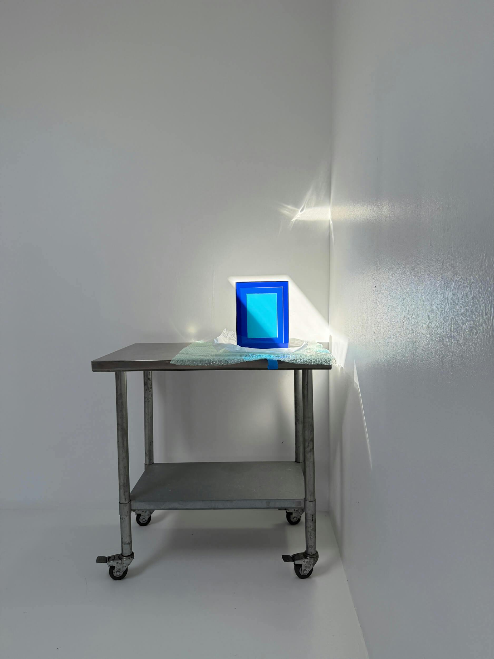 A small metal cart stands against white walls. On top, a geometric blue artwork is illuminated by sunlight, casting sharp shadows and reflections on the wall. The cart has two shelves and wheels.