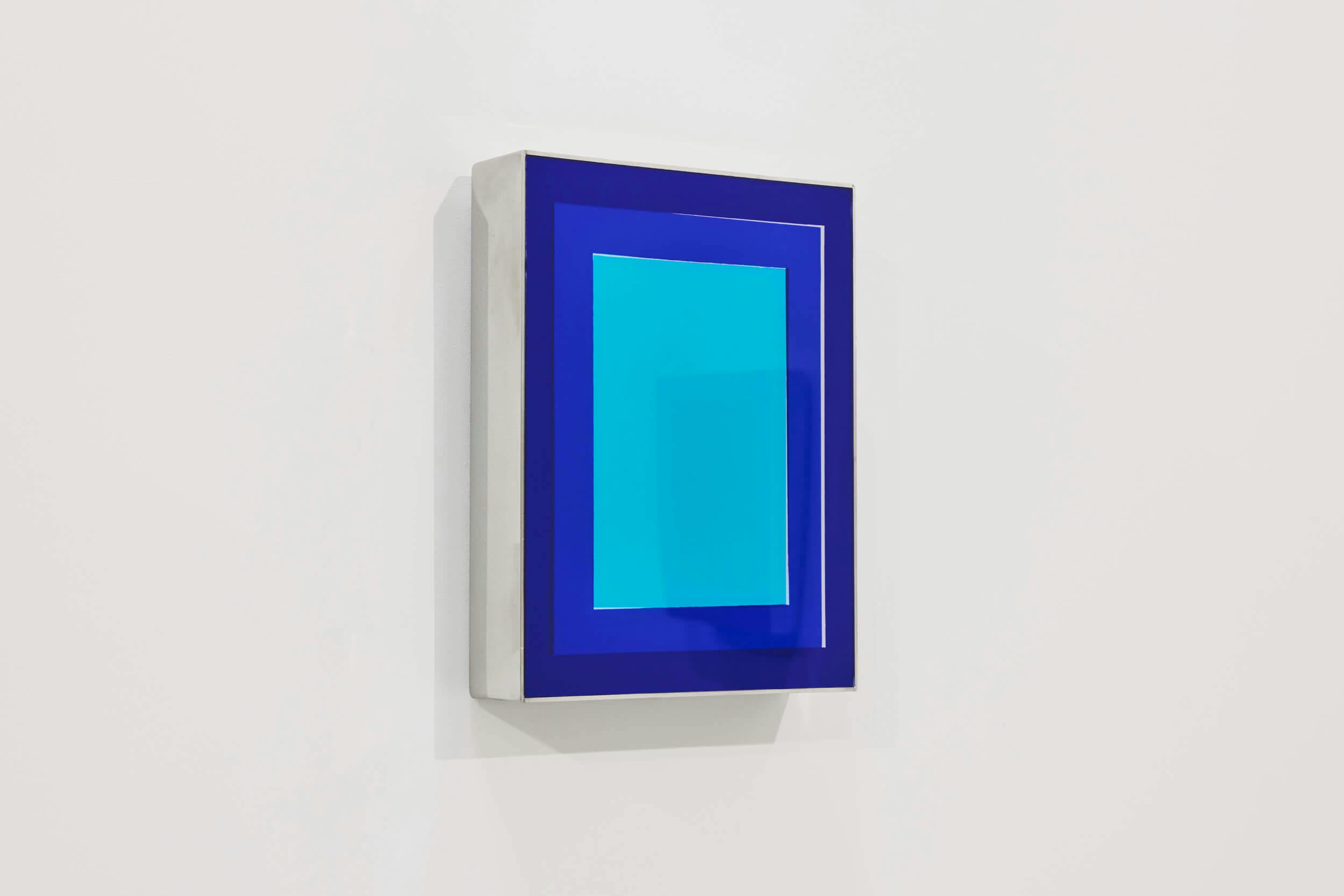 A rectangular, modern art piece with a layered design, featuring a light blue center bordered by darker blue and navy blue rectangles, mounted on a white wall. The frame appears slightly three-dimensional.