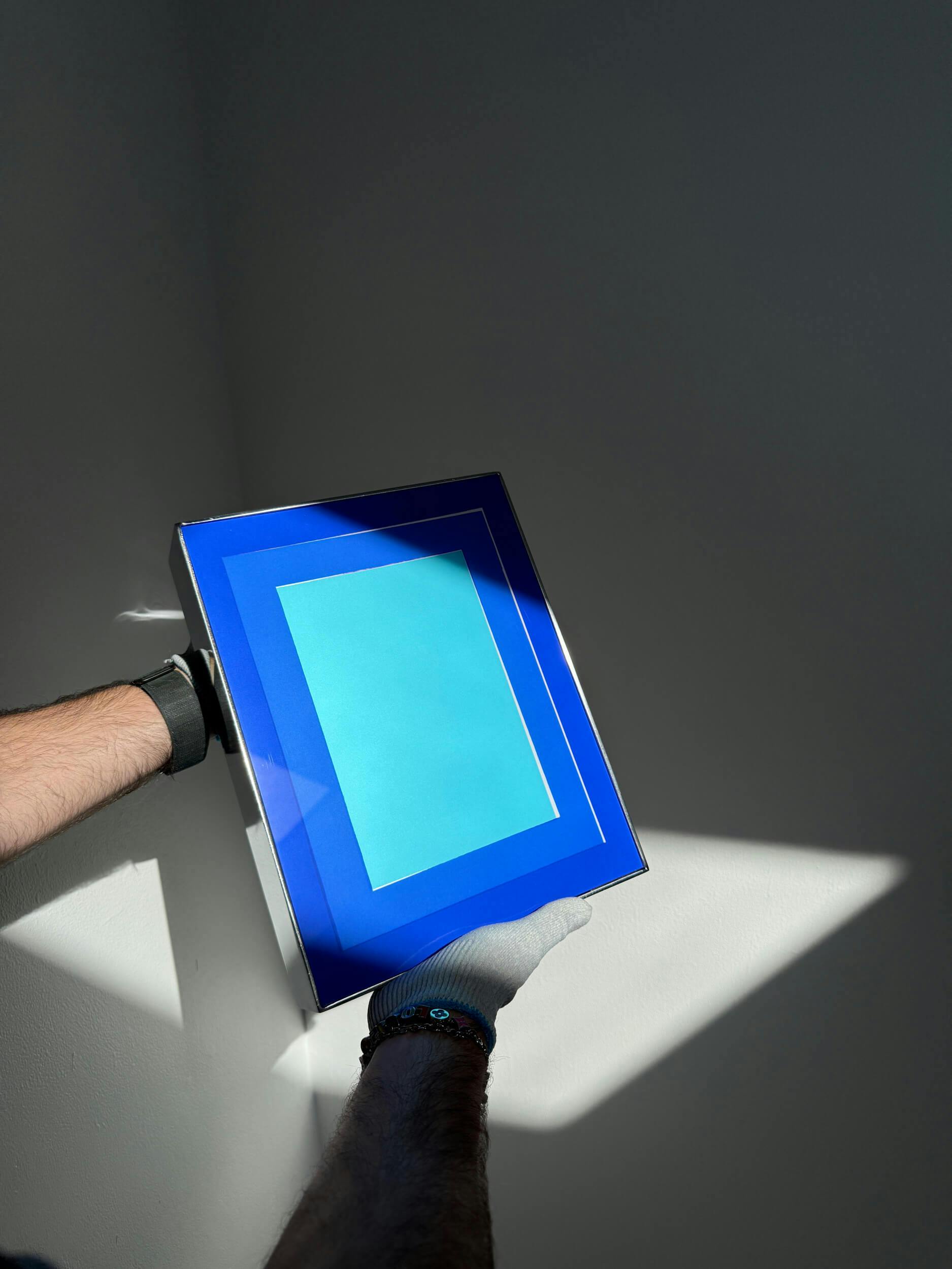 A person wearing gloves holds a framed artwork with light blue and dark blue rectangular shapes, standing in a patch of sunlight against a plain gray wall.