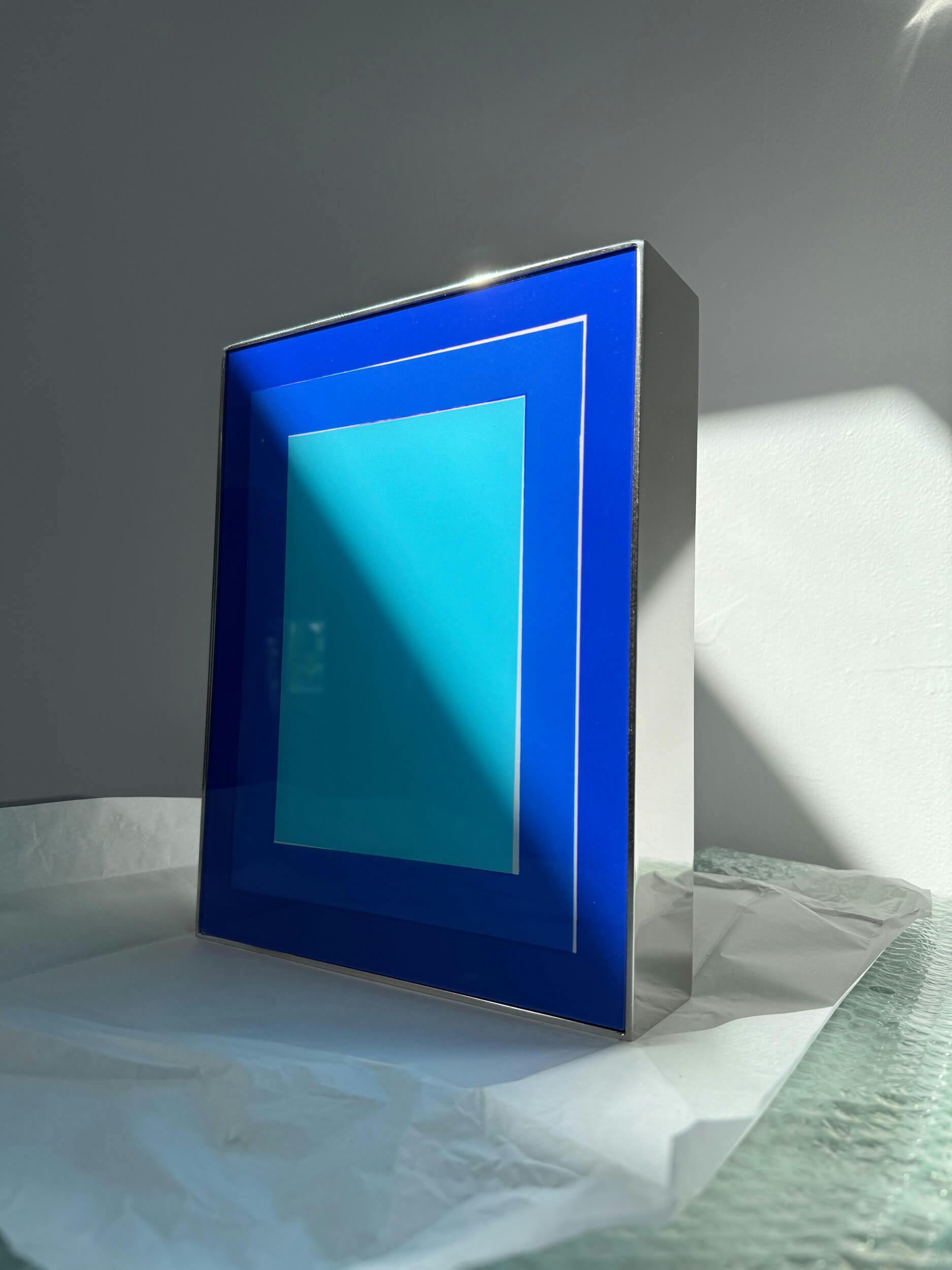 A rectangular object with a shiny metallic frame and a blue geometric design stands upright on a textured glass surface, illuminated by sunlight coming from the right side. White paper is partially visible underneath.