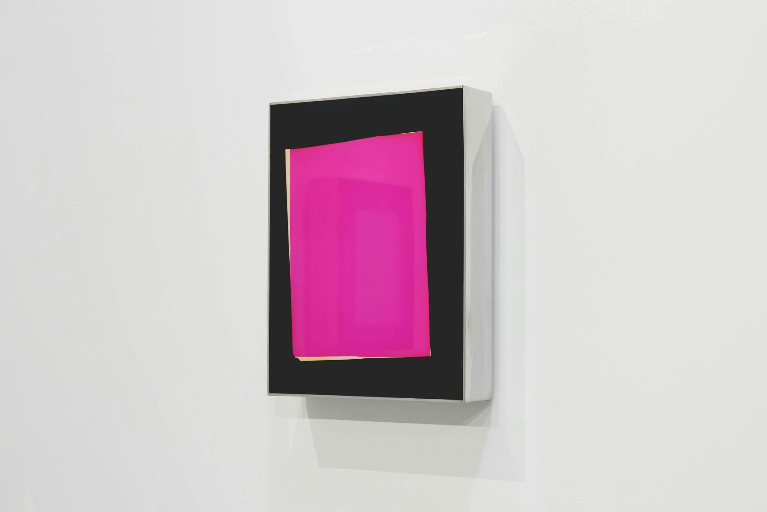 A rectangular artwork with a black border displaying a bright pink, semi-transparent geometric shape is mounted on a white wall. The shape appears layered and slightly off-center within the frame.