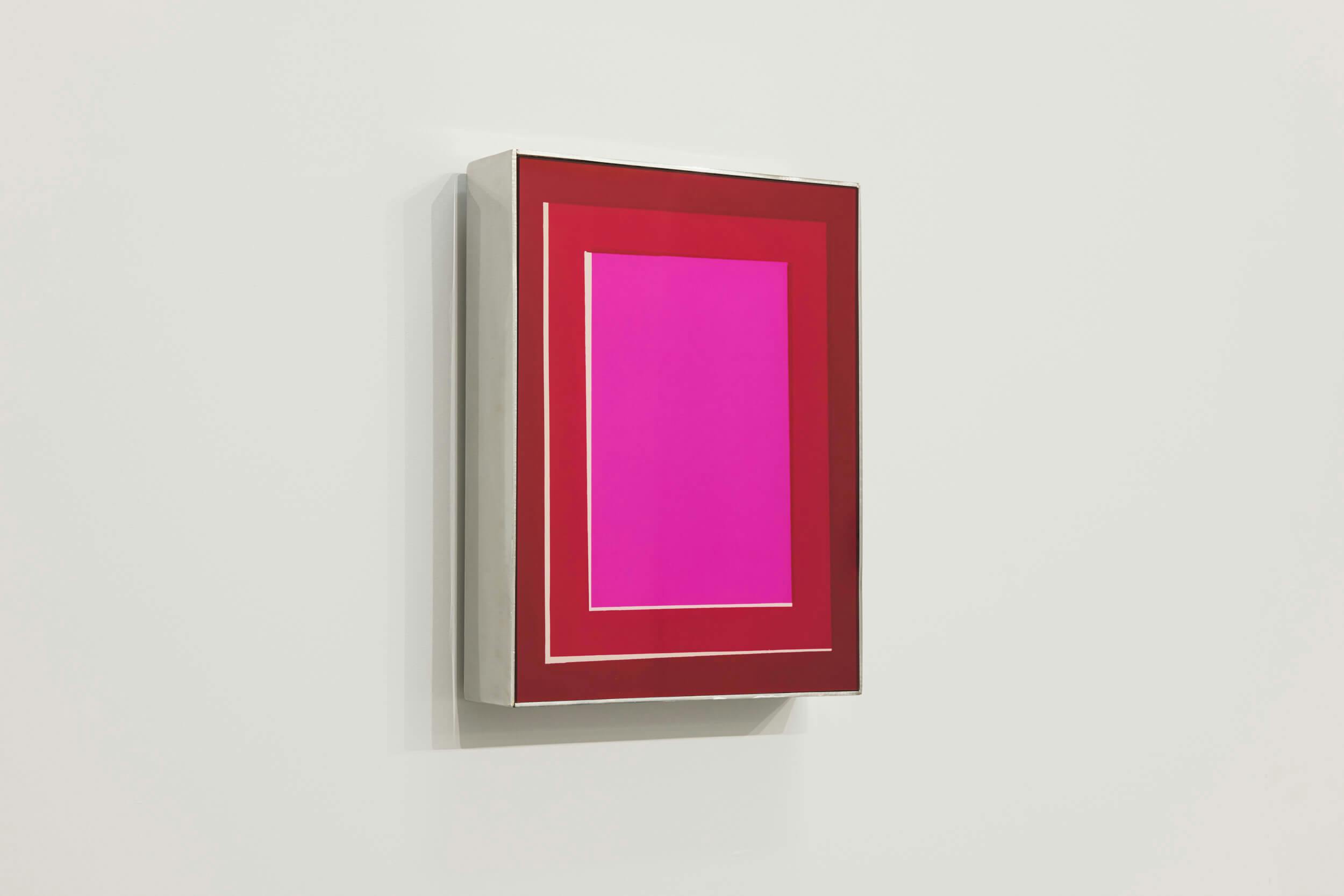 A rectangular artwork with a bright pink center, surrounded by red and white rectangular borders, hangs on a white wall. The frame extends outward, giving the piece a three-dimensional appearance.