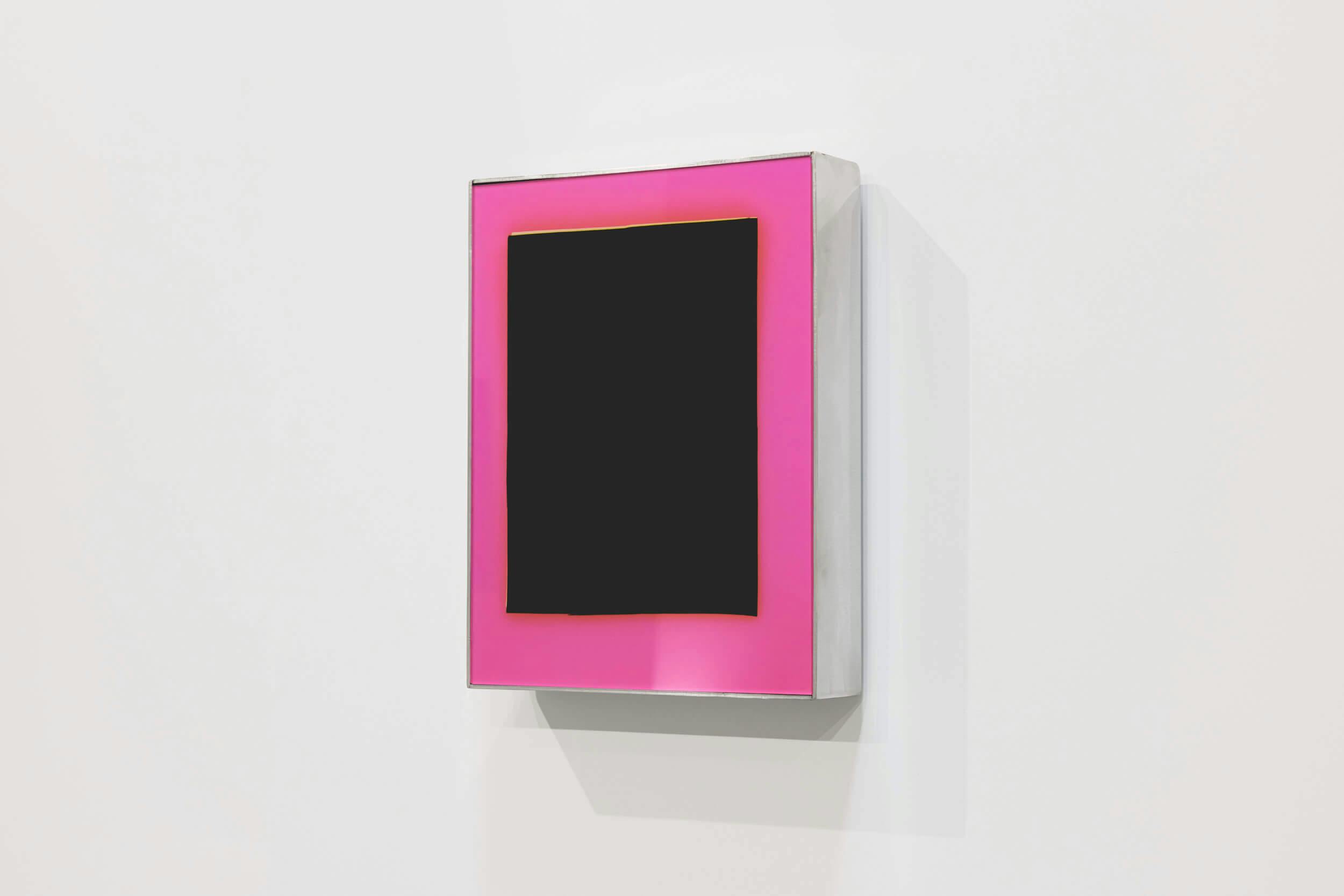A rectangular black panel is centered within a bright pink border, mounted on a white wall. The artwork has a three-dimensional, box-like appearance and casts a faint shadow on the wall.
