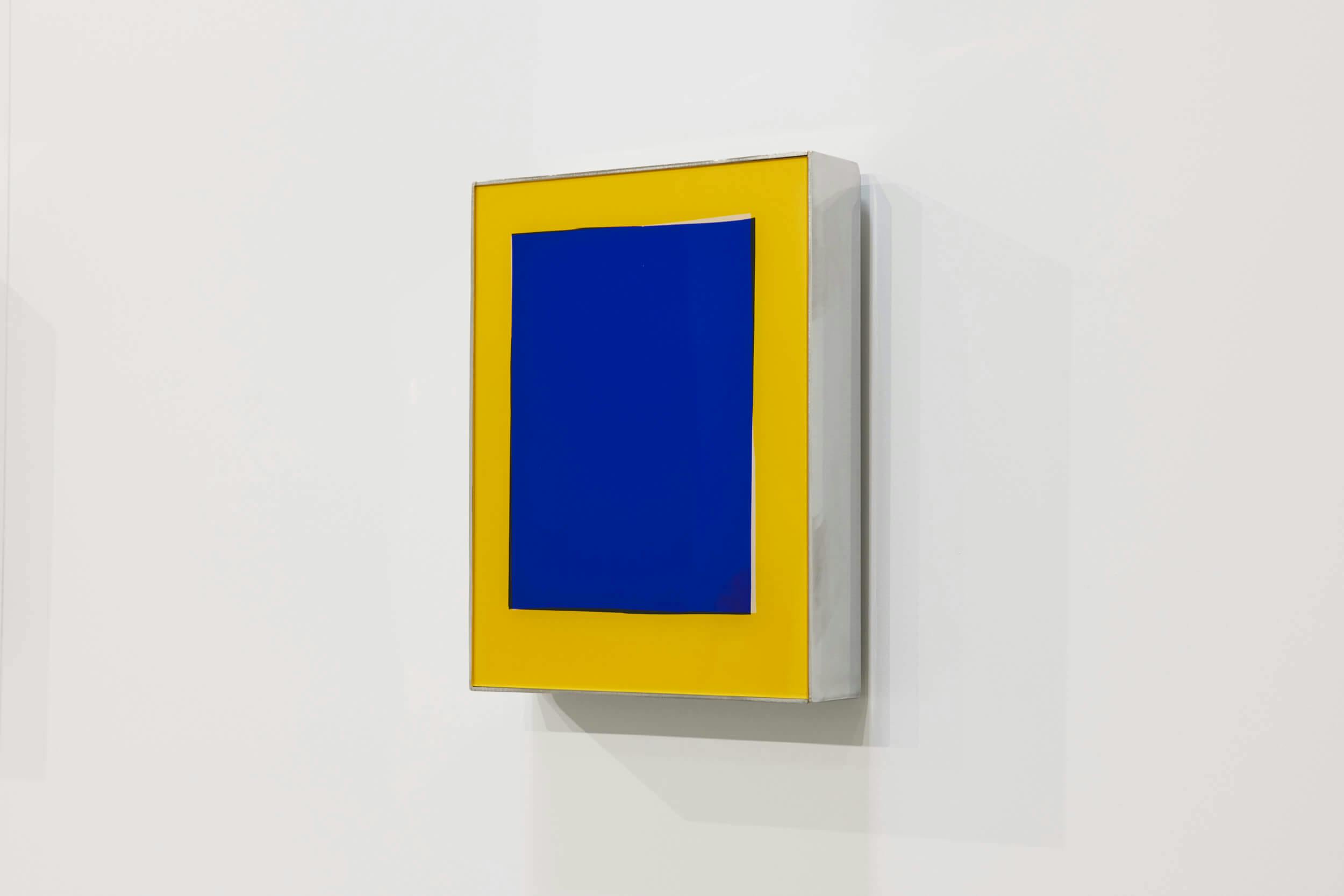 A rectangular wall-mounted artwork features a blue rectangle centered on a yellow background, both enclosed in a white, box-like frame against a plain white wall.