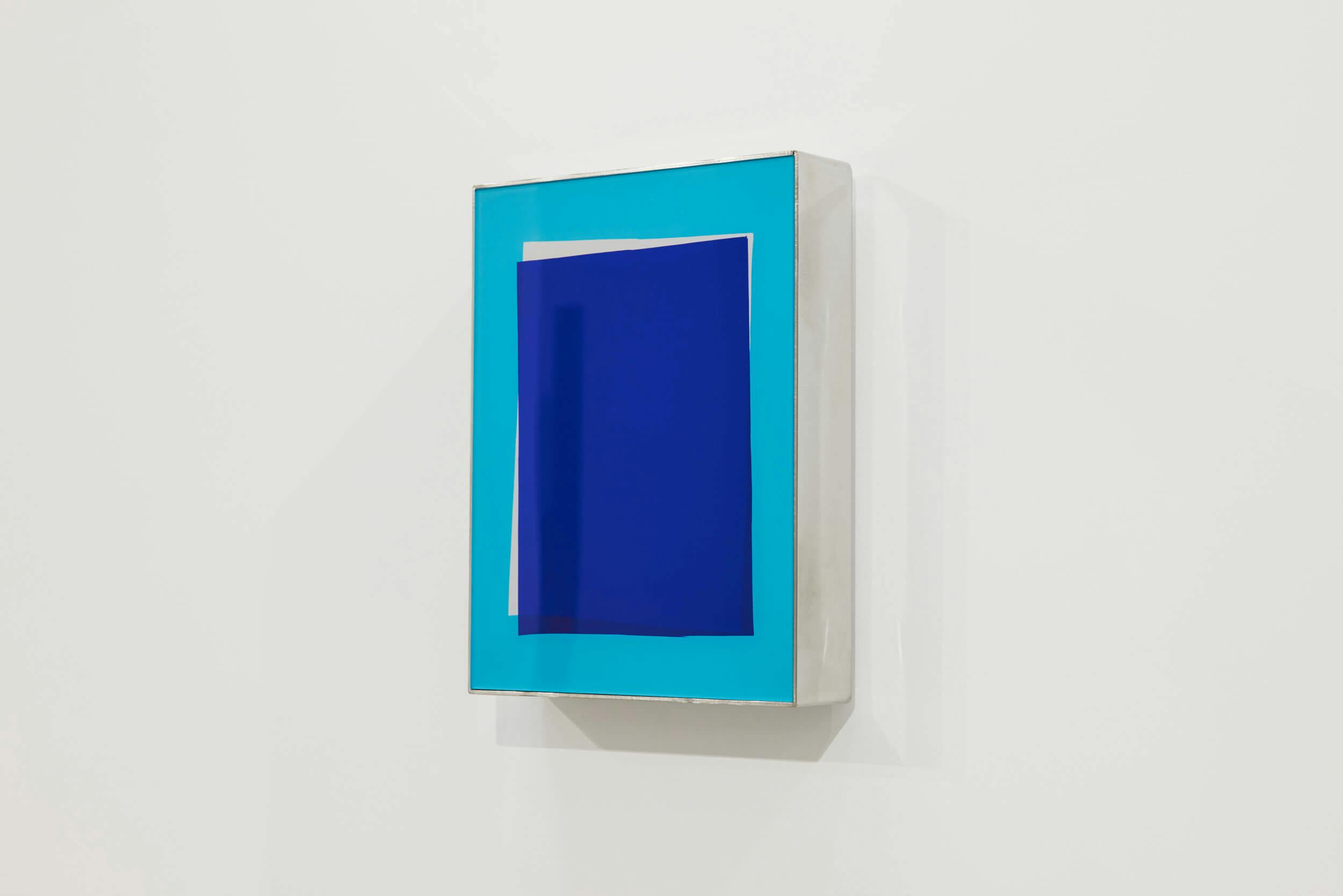 A rectangular artwork with a blue and turquoise geometric design hangs on a white wall. The piece consists of layered rectangles in varying shades of blue within a clear frame.