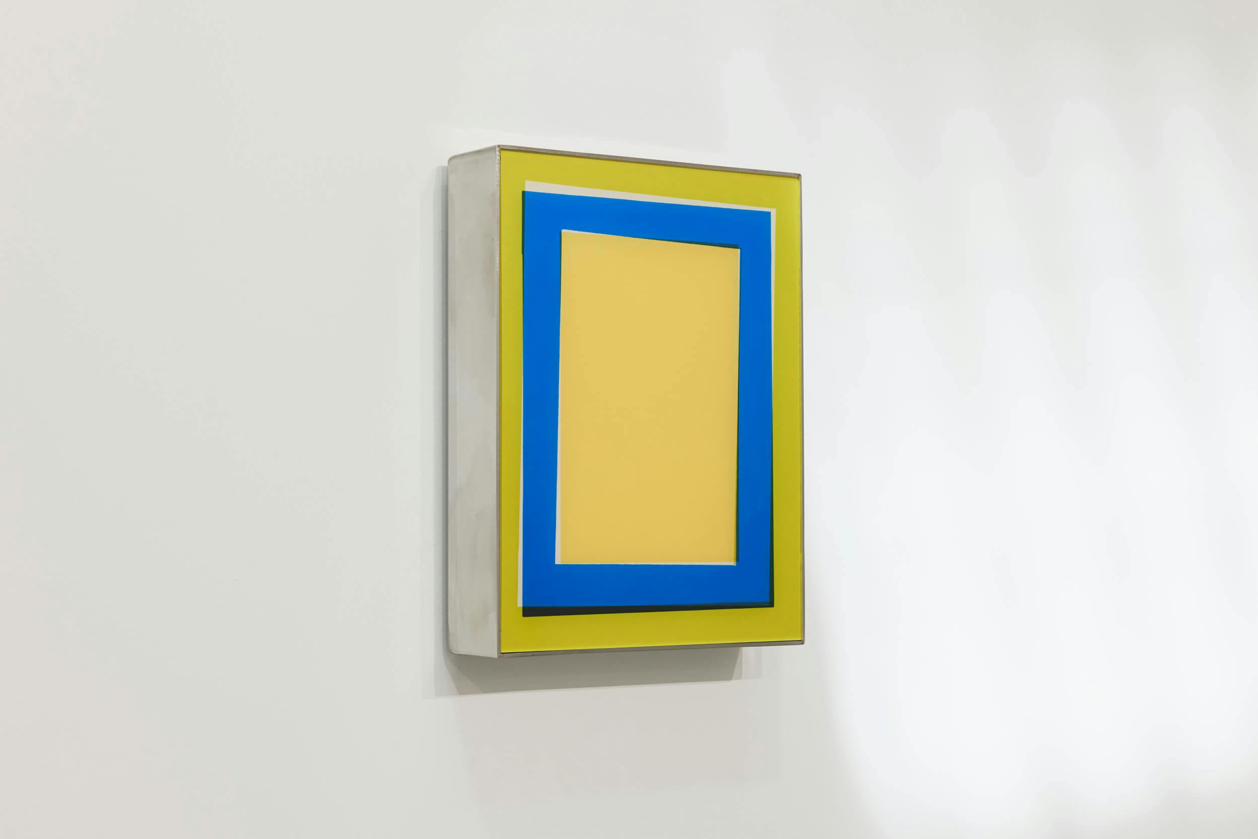 A rectangular artwork mounted on a white wall features a yellow background bordered by a thick blue frame, with an additional yellow border outside the blue. The piece is displayed in a silver or gray case.