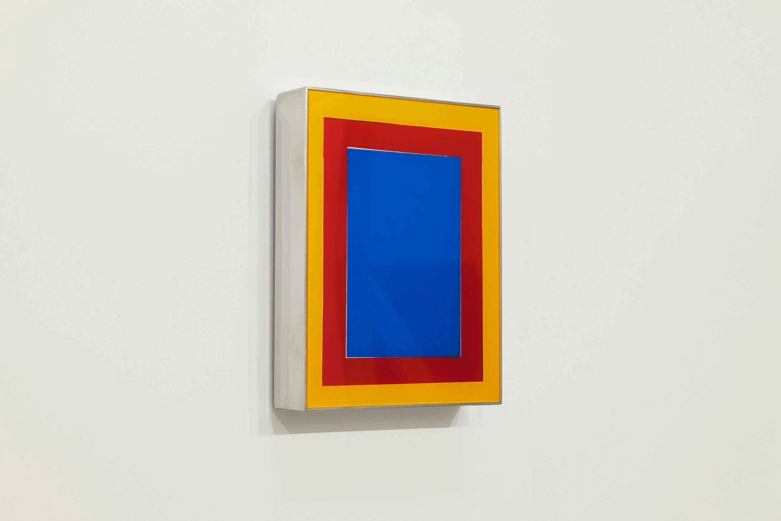 A rectangular artwork with concentric colored borders—blue in the center, surrounded by red and orange—mounted on a white wall. The piece appears three-dimensional with a reflective frame.