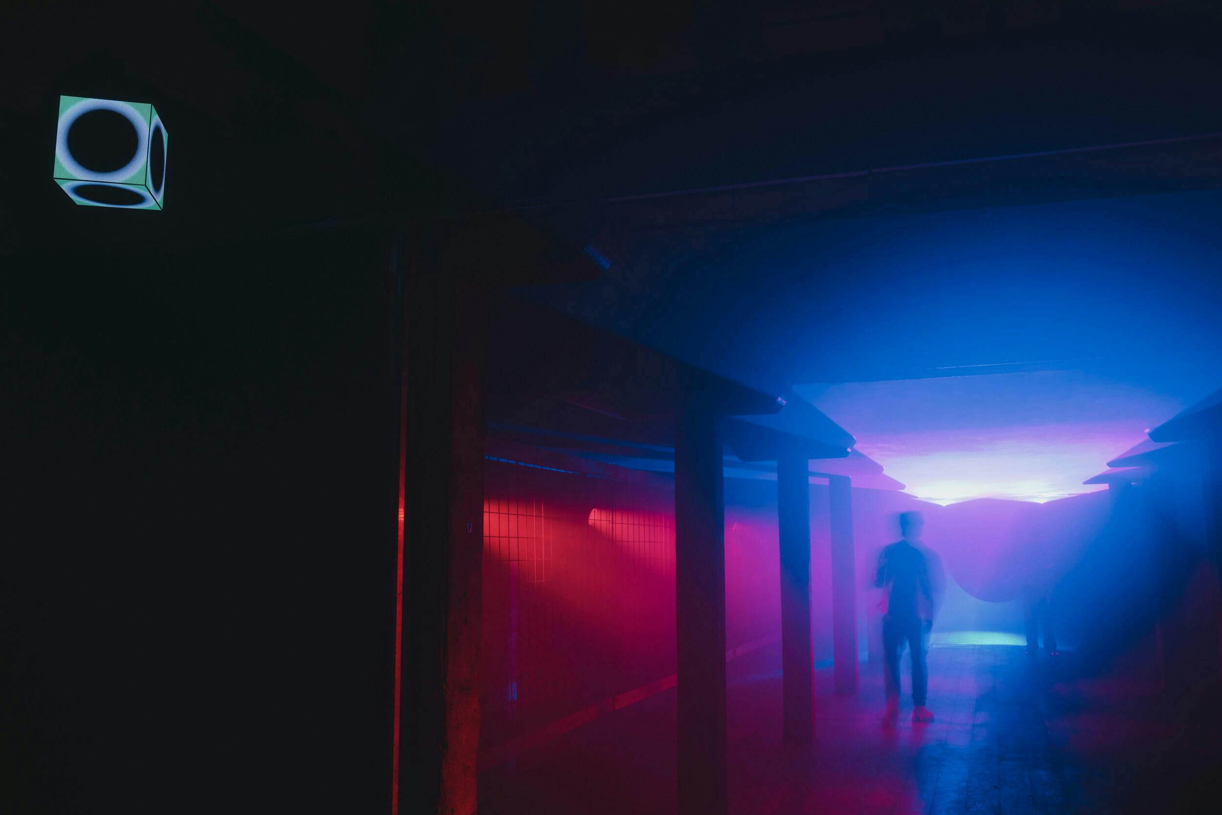 A person walks through a dimly lit hallway with blue and pink lights, shadowy pillars, and a glowing cube suspended from the ceiling in the upper left corner.