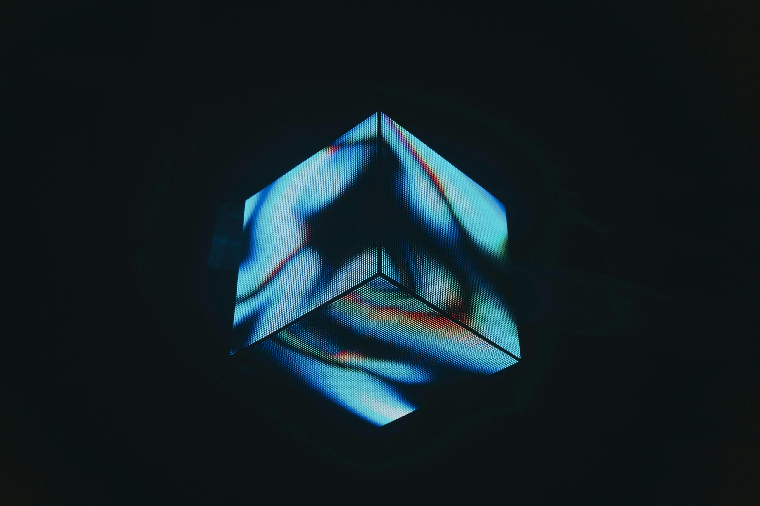 A geometric cube with illuminated, abstract, blue and white swirling patterns is suspended against a dark background. The lighting creates a futuristic or digital effect.