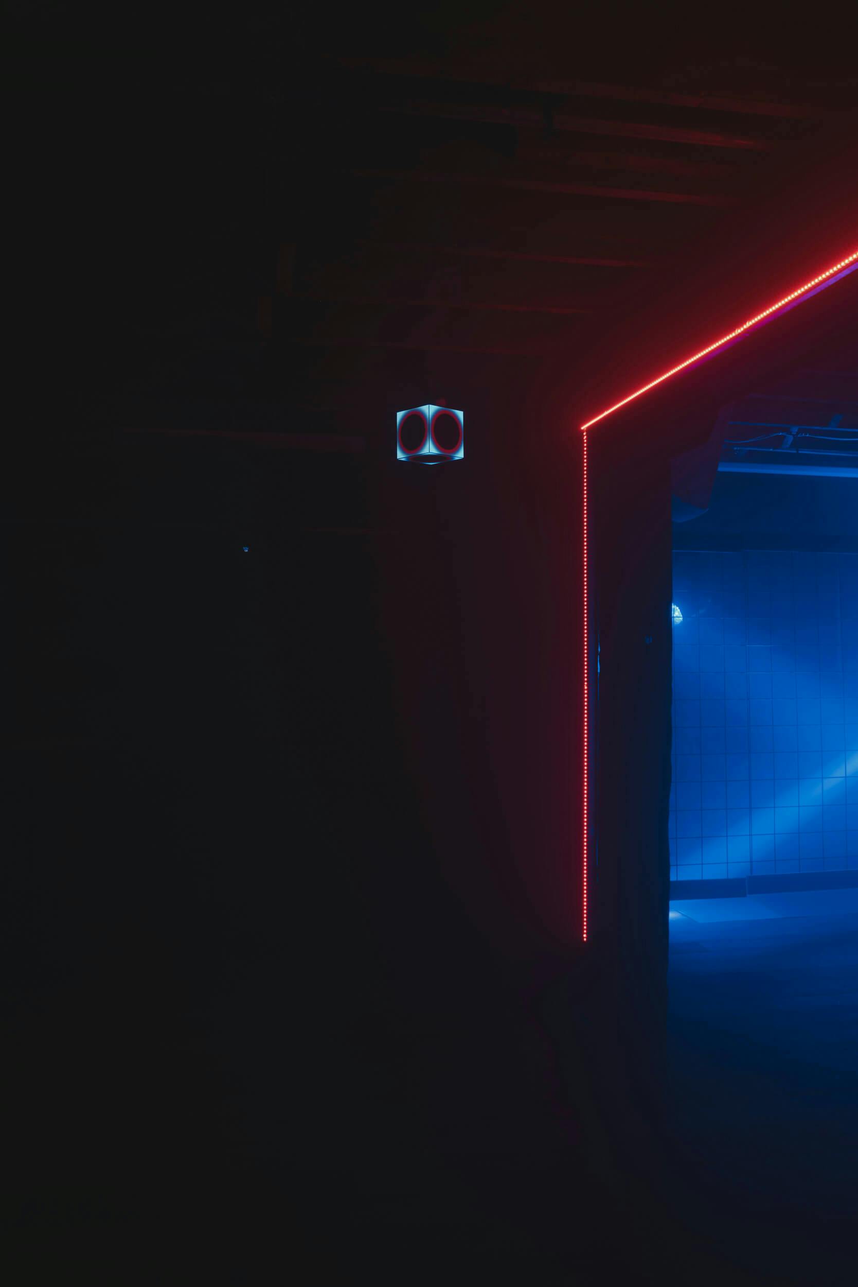 A dark room with a corner outlined by red neon lights, partially revealing a blue-lit tiled wall in the background and a small security camera mounted near the ceiling.