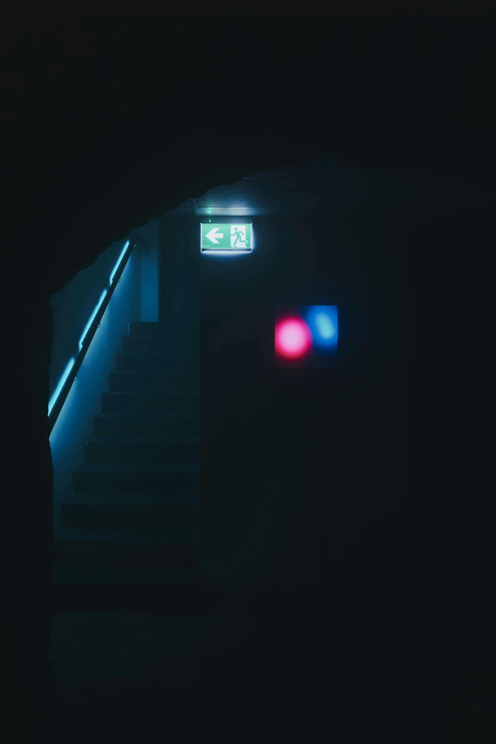 A dimly lit stairwell with a blue handrail, an illuminated green exit sign above the stairs, and a faint reflection of red and blue light on a nearby wall. The surrounding area is mostly dark.