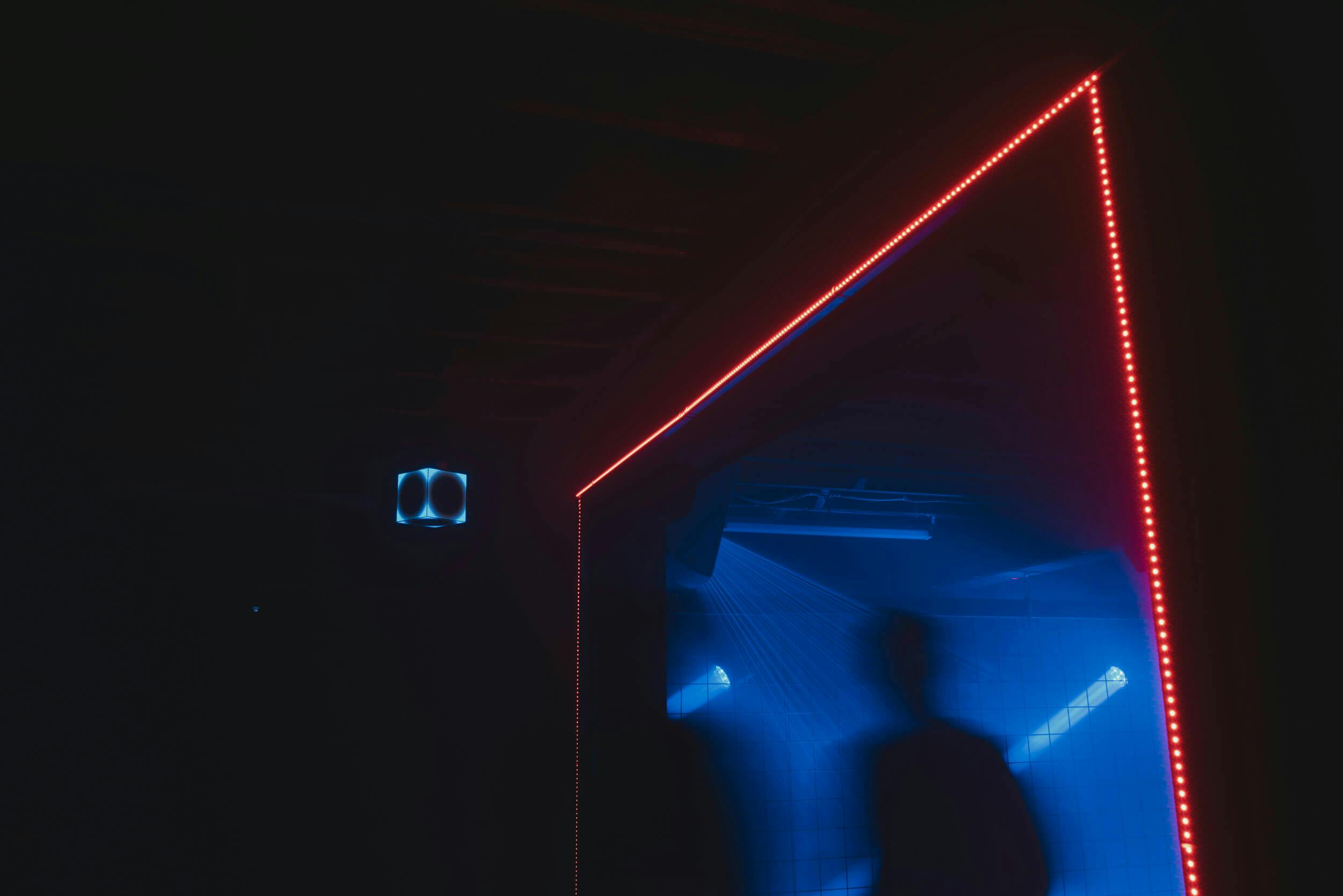 A dimly lit room with blue lighting, two blurred human figures, a line of red LED lights framing a doorway, and geometric shapes illuminated on the wall.