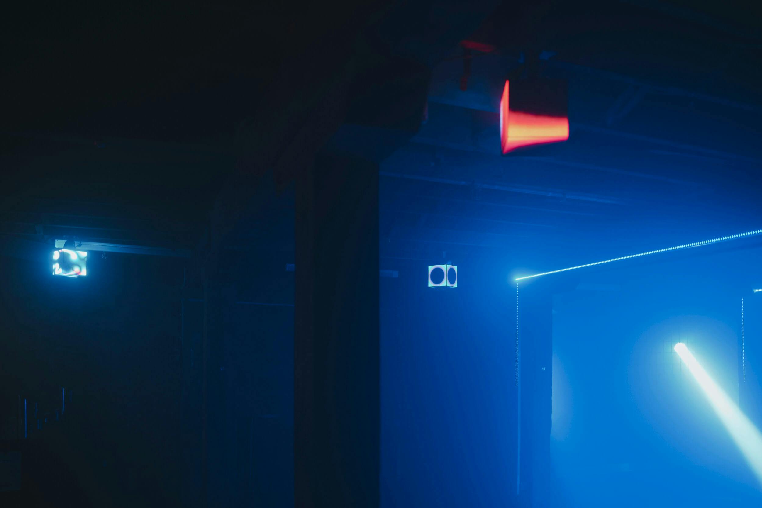 A dimly lit room with a blue light illuminating part of the space. A red exit sign hangs from the ceiling, and beams of light create geometric shapes in the foggy atmosphere.