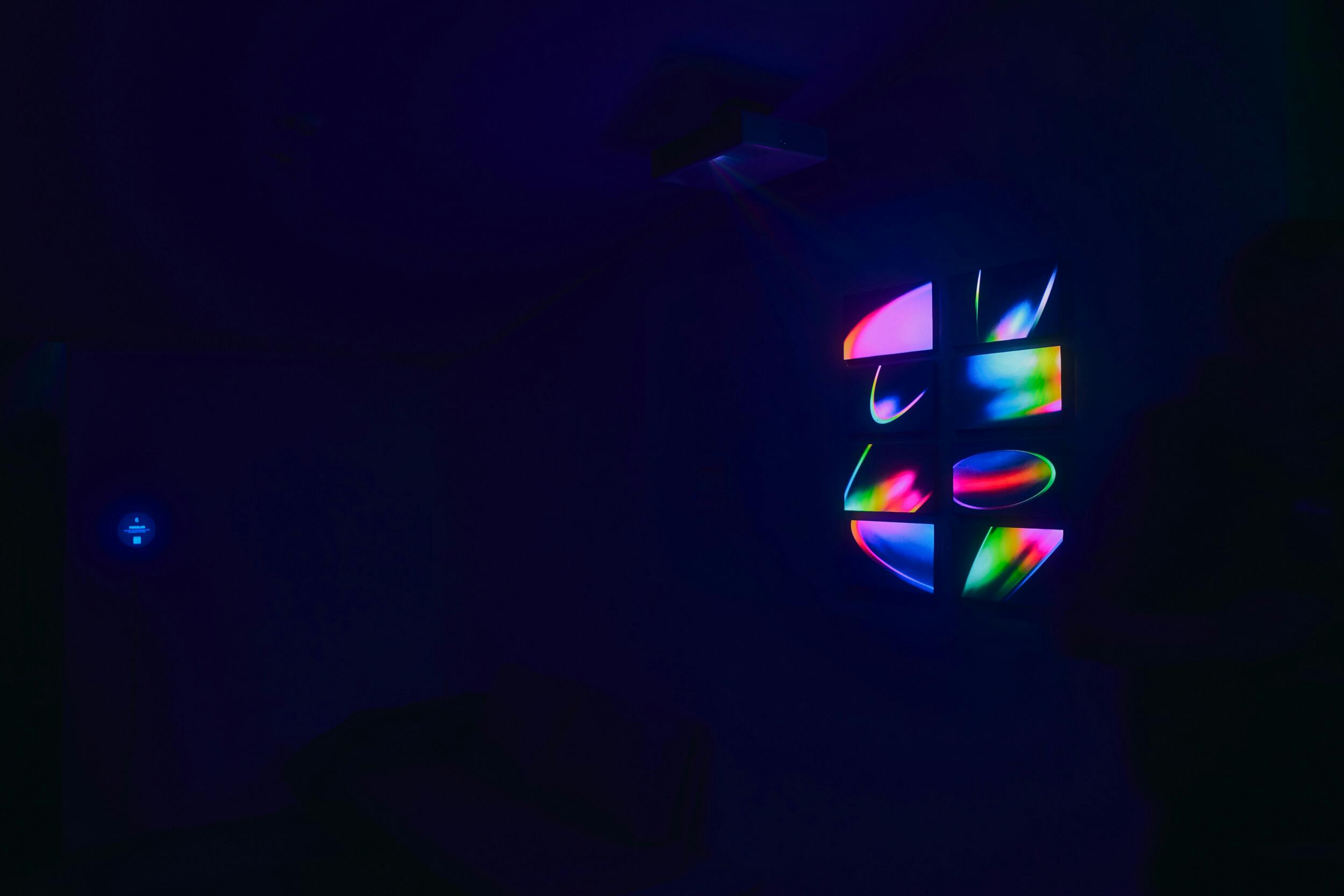 A dark room with a colorful, abstract, and segmented light projection on the wall, consisting of curved and geometric shapes. A faint blue circle of light appears on the opposite wall.