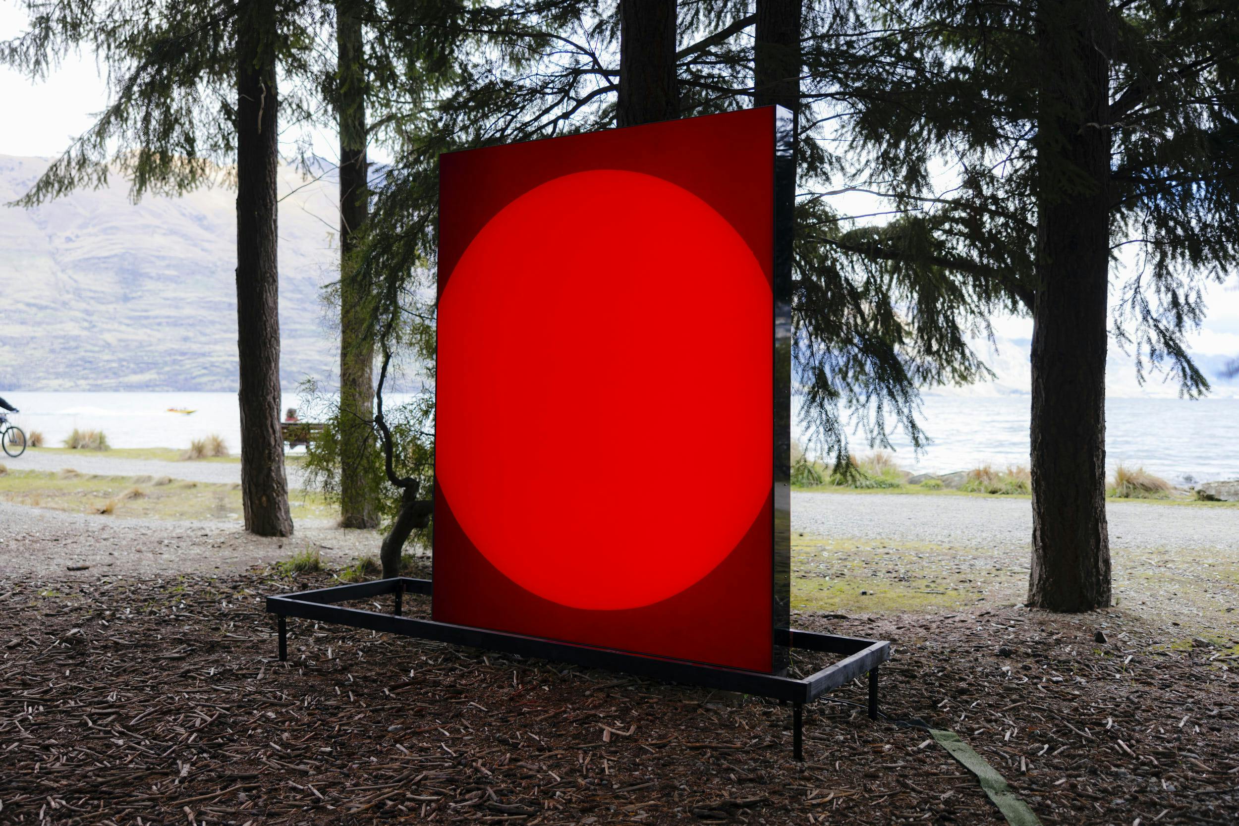 A large rectangular panel with a bright red circle in the center stands among trees near a lake, with water and mountains visible in the background.