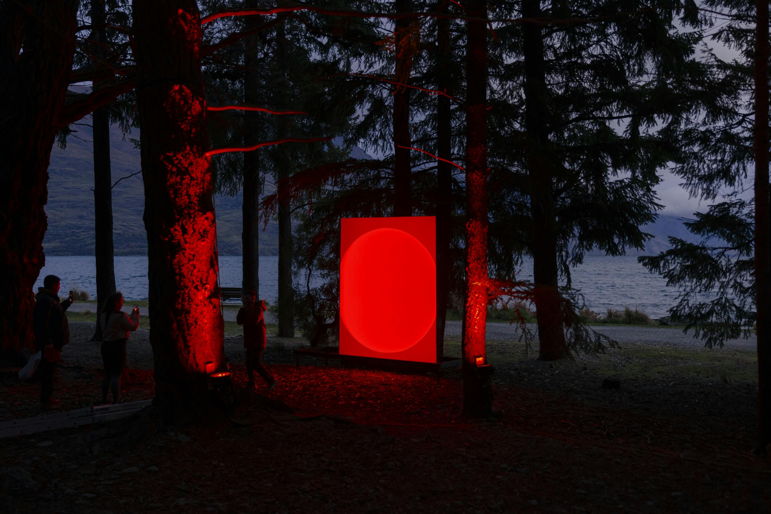 A large glowing red rectangular panel stands among trees near a lake at dusk, with red lights illuminating the nearby tree trunks and several people standing around the installation.
