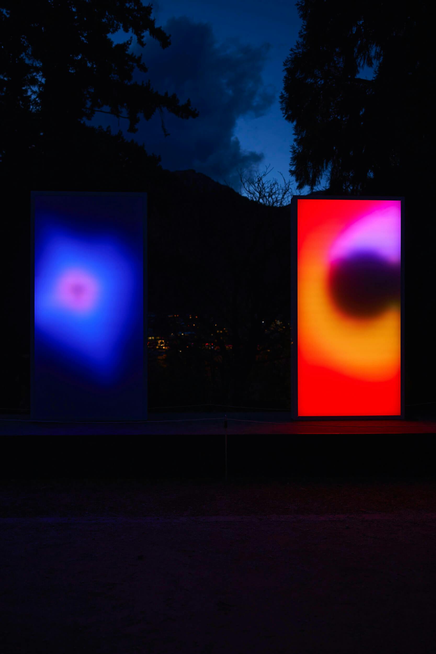 Two tall rectangular screens stand outdoors at night; the left screen displays a blue and white gradient, while the right shows a vibrant gradient of red, yellow, orange, and black. Trees and mountains are visible in the background.