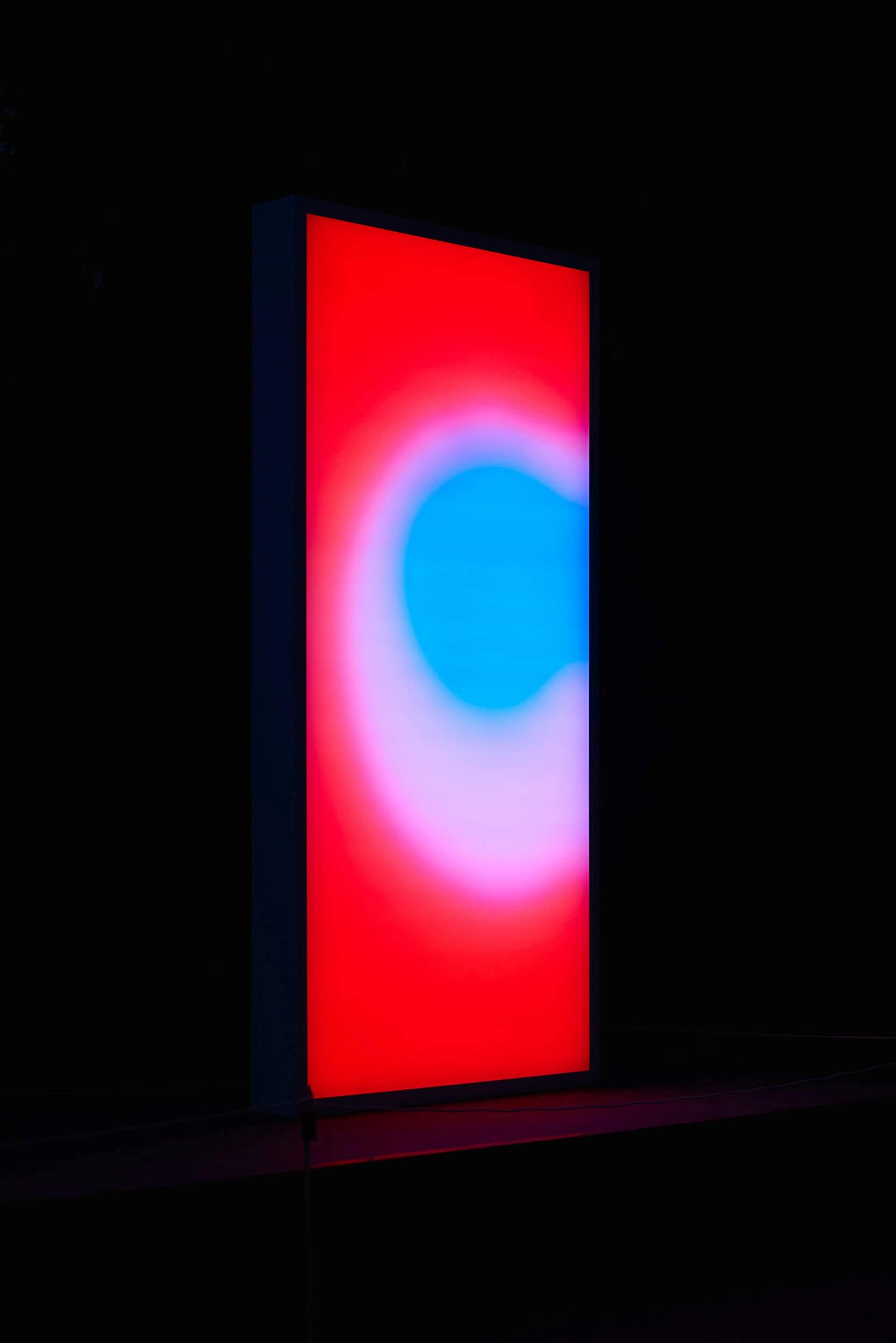A vertical rectangular screen displays a bright abstract pattern with a blue circle in the center, surrounded by pink and red gradients, against a dark background.