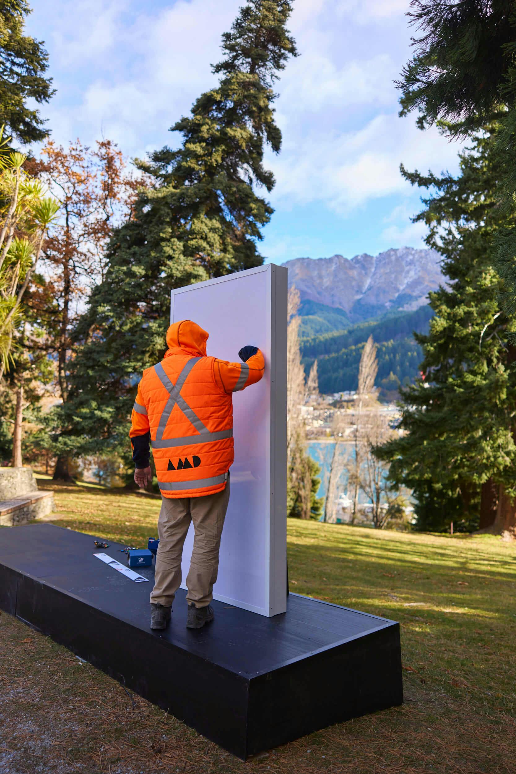 A person in an orange high-visibility jacket stands on a black platform outdoors, working on a large vertical panel with trees, grass, and mountains in the background.