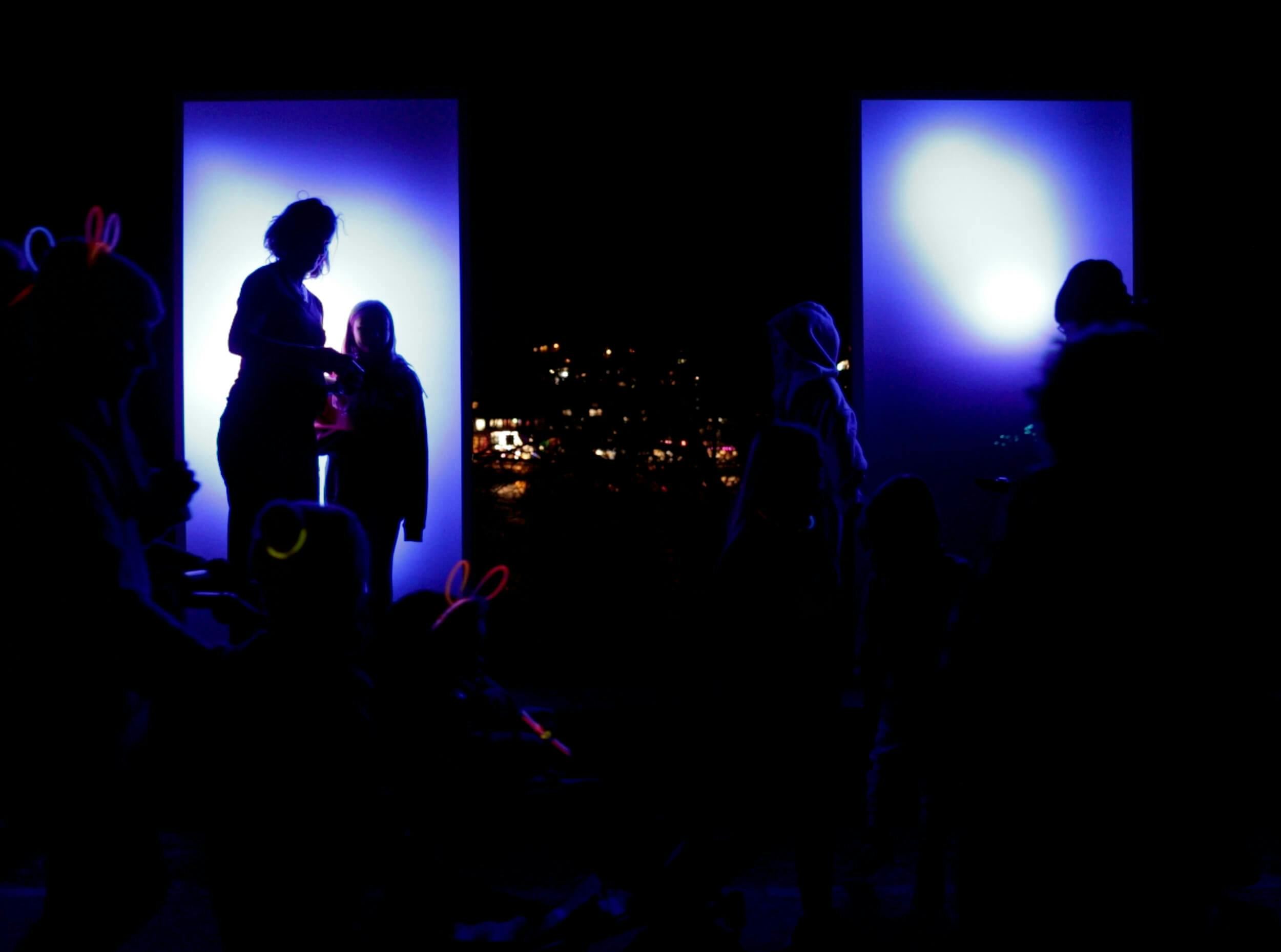 A group of people, mostly in silhouette, stand and interact in front of two large, illuminated blue panels at night. Some are wearing glow sticks and bunny ear headbands. City lights are visible in the background.