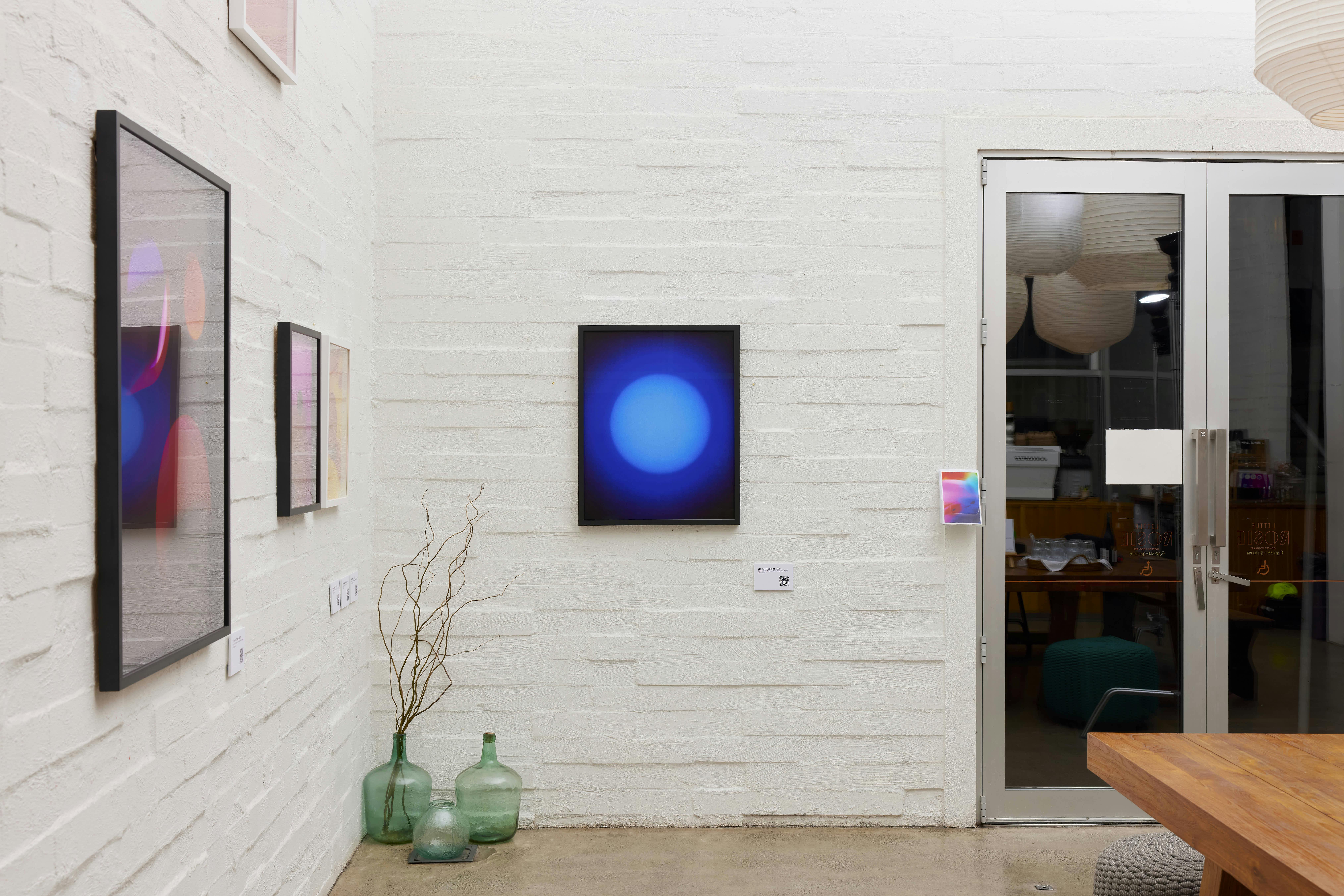 A modern art gallery with white brick walls displays several framed abstract artworks. Three large green glass bottles and a branch sit in a corner. Glass doors on the right reveal a glimpse of another room.