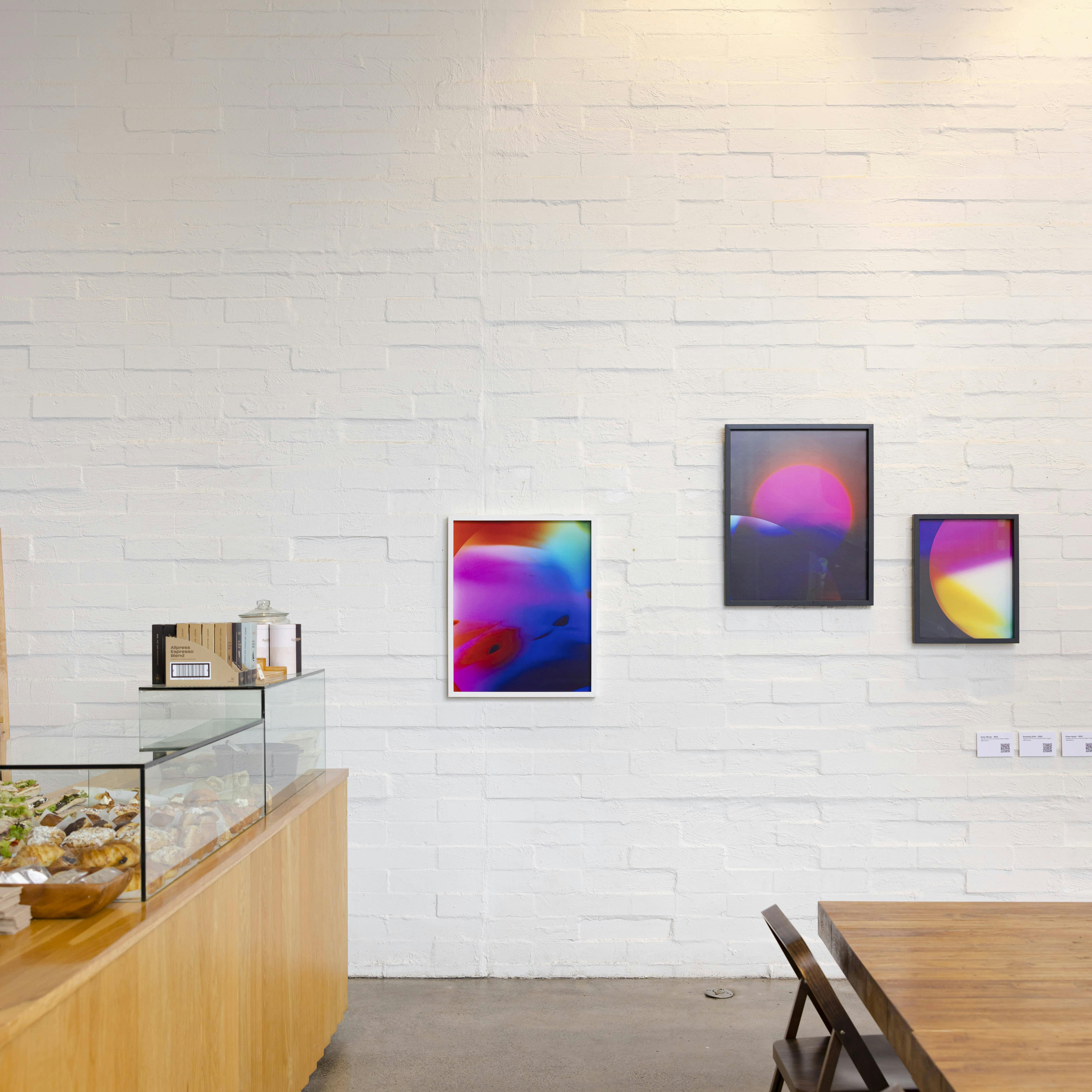 A modern cafe interior with a glass display of pastries on the left and three framed abstract artworks with bright colors hanging on a white brick wall above a wooden table and chairs.