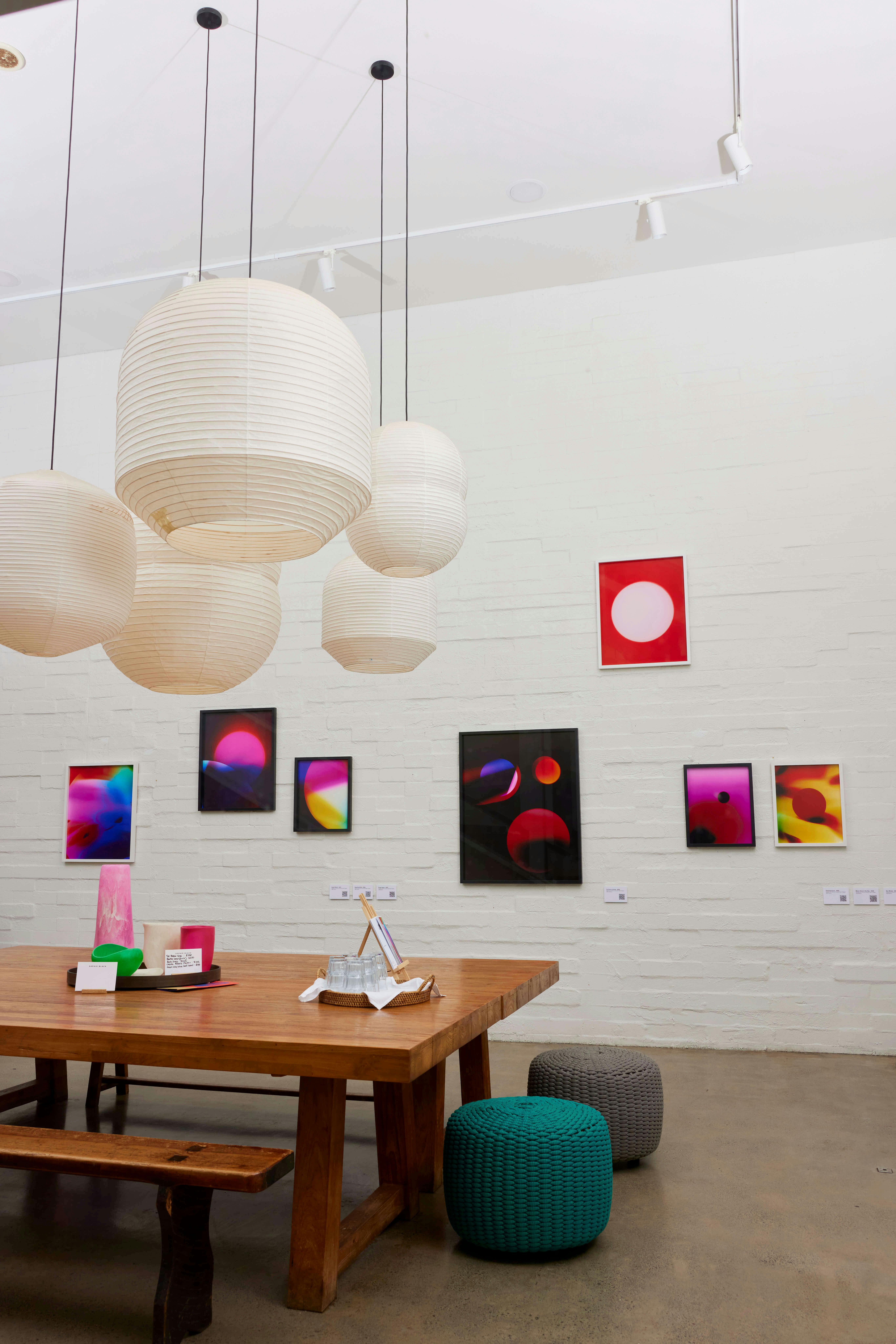 A modern room with a wooden table, paper lantern pendant lights, and colorful abstract artwork on a white brick wall. Round knitted ottomans and a concrete floor are also visible.