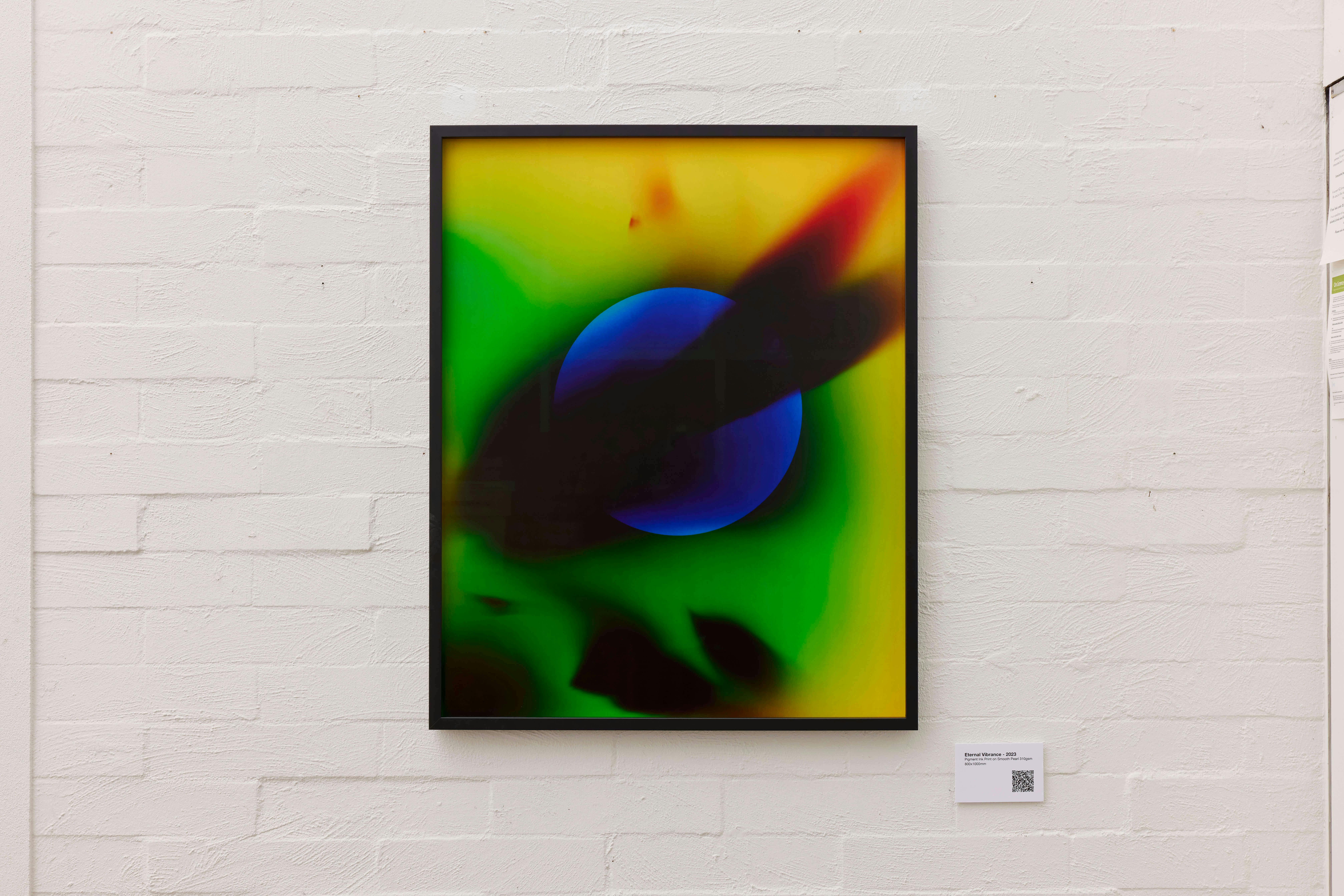 A framed abstract artwork features a blue sphere at the center with green, yellow, and red gradients surrounding it, hanging on a white brick wall with a small label and QR code displayed nearby.
