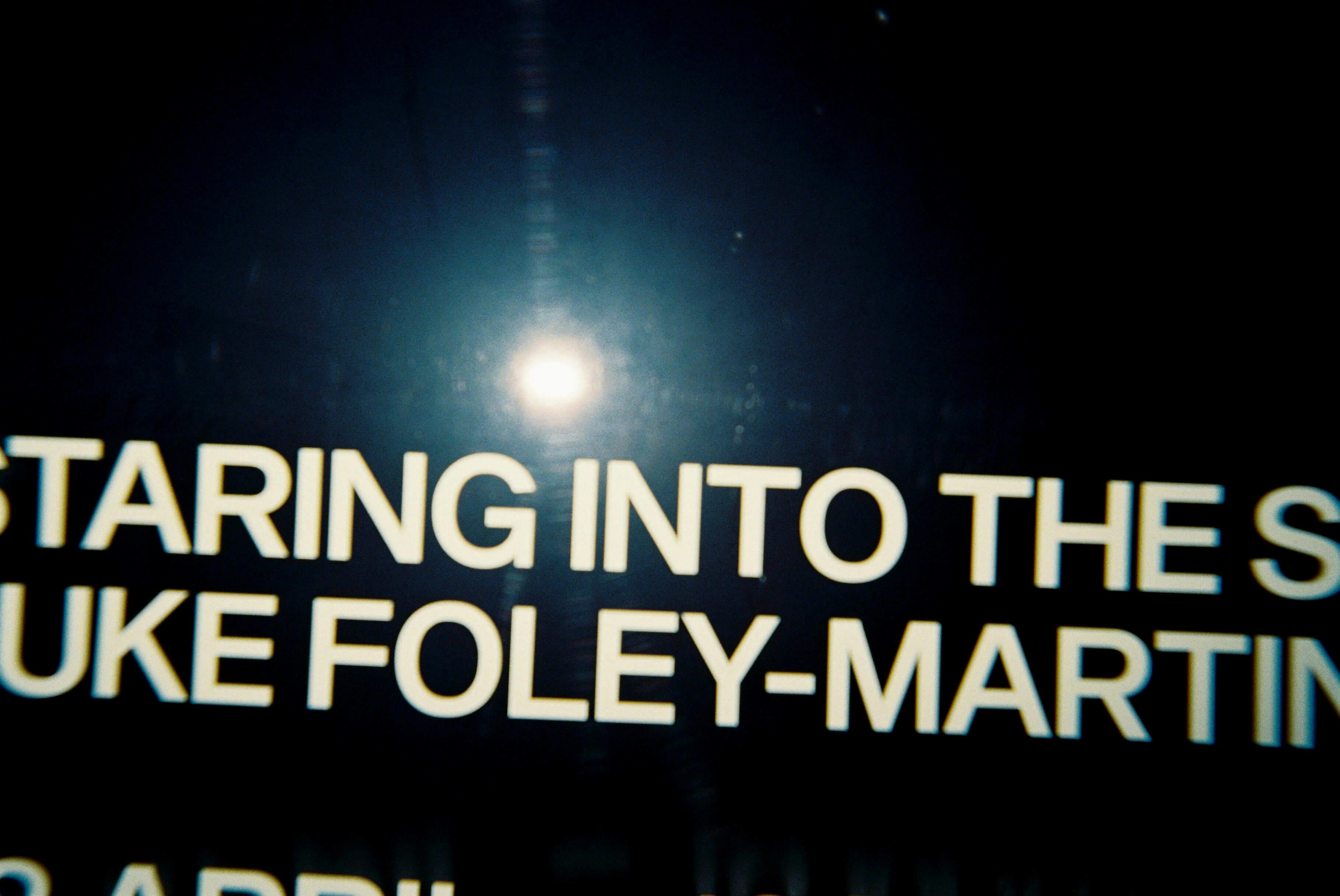 A close-up photo of white text on a dark background, partially reading “STARING INTO THE S” and “UKE FOLEY-MART,” with a bright light reflection obscuring part of the text.