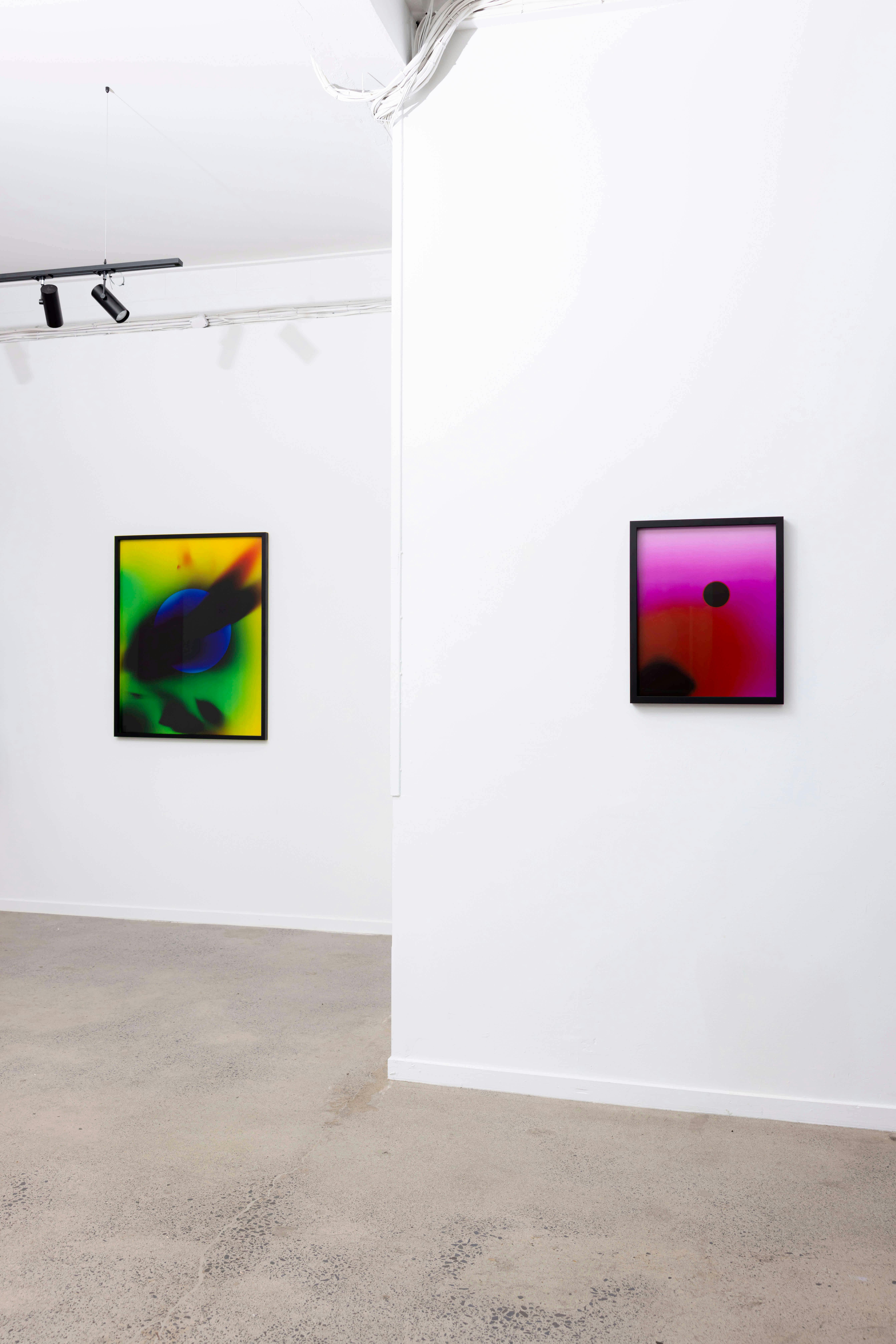 A white-walled art gallery displays two abstract framed prints. The left print has green, yellow, and purple tones; the right print features red and purple shades with a black dot. The floor is light-colored concrete.