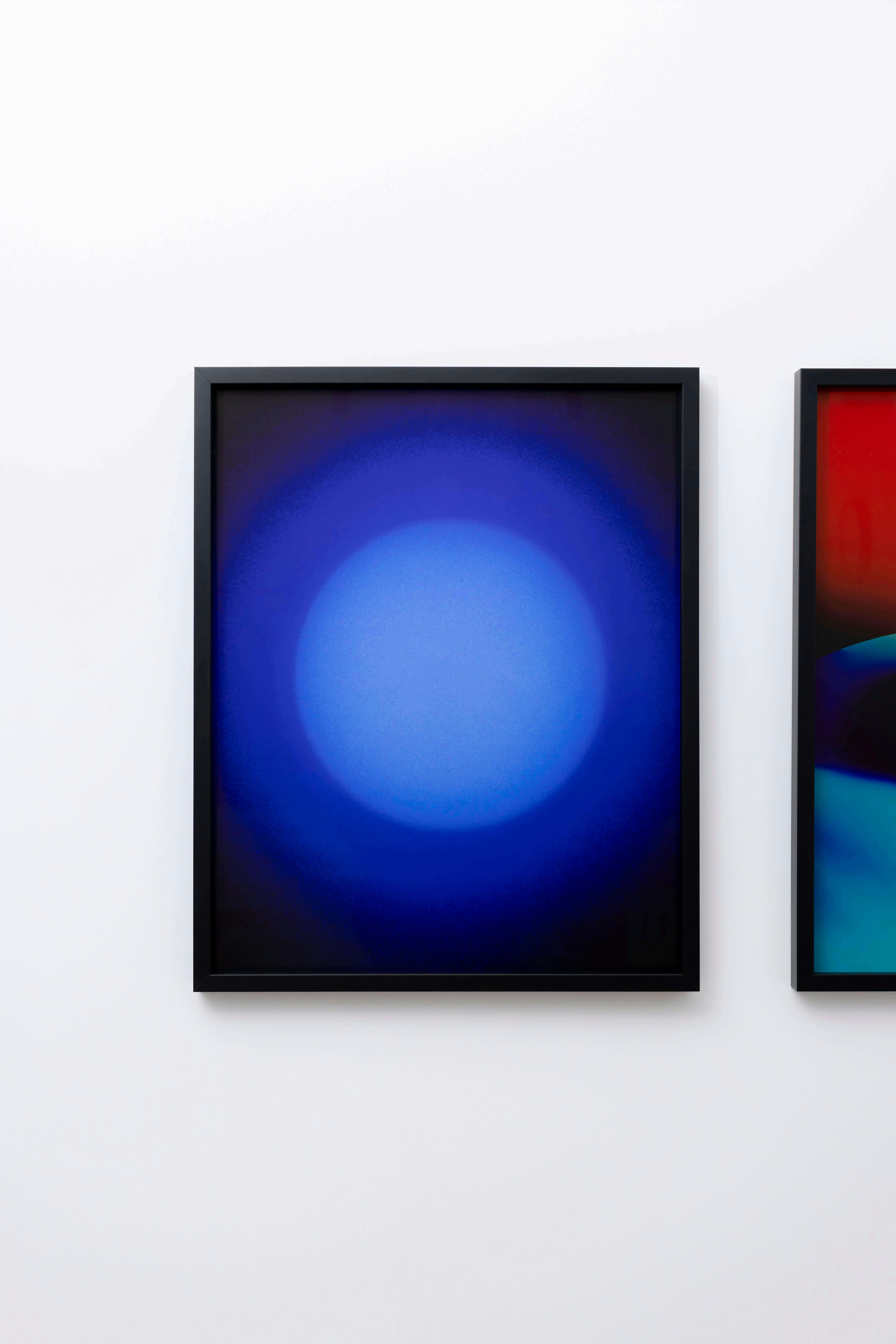 A framed abstract artwork featuring a large, glowing blue circle with a soft gradient against a darker blue background, hung on a white wall. A portion of another colorful artwork is visible to the right.