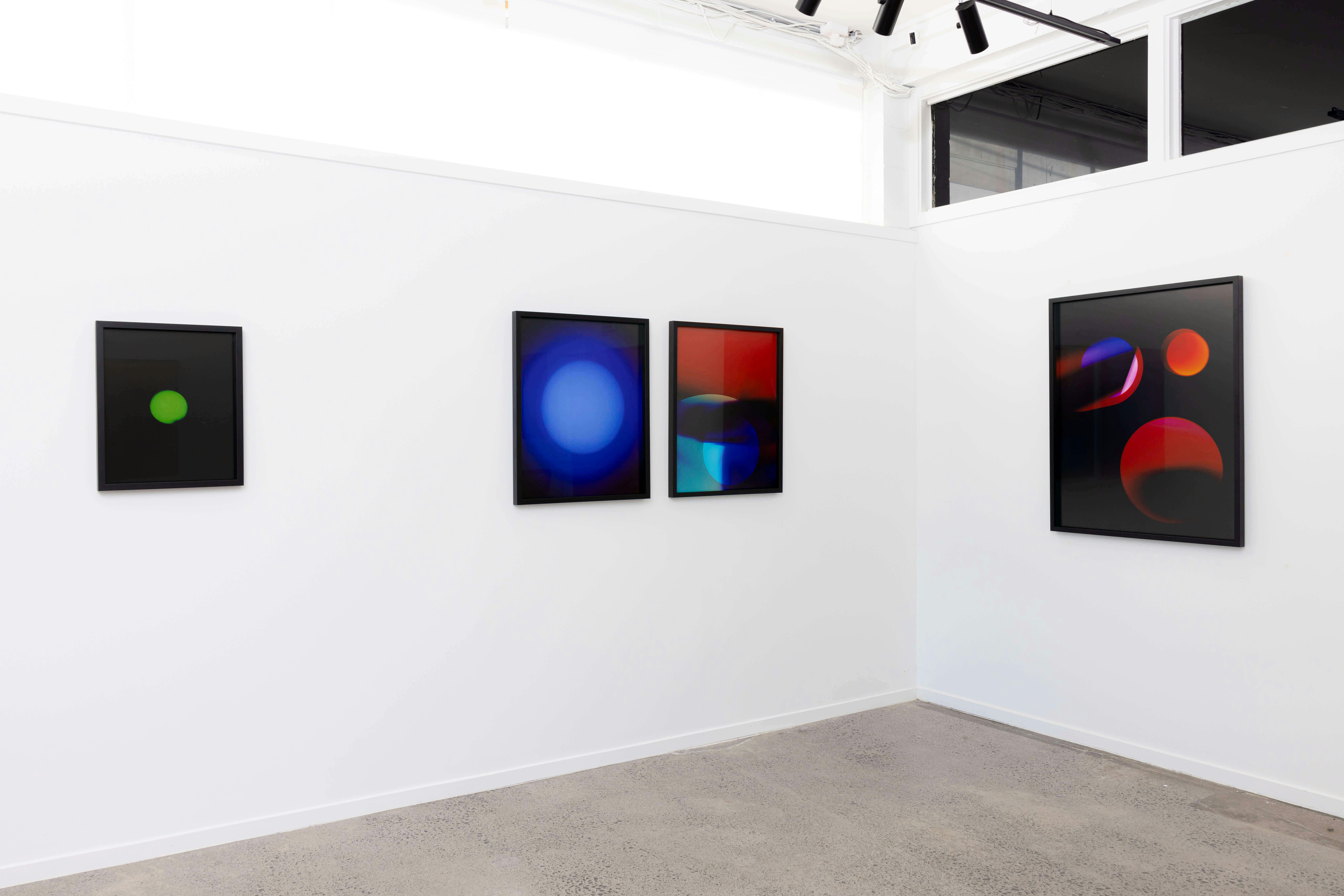 A white gallery wall displays four framed abstract artworks featuring colorful, glowing circular shapes on dark backgrounds. The gallery has a concrete floor and track lighting on the ceiling.