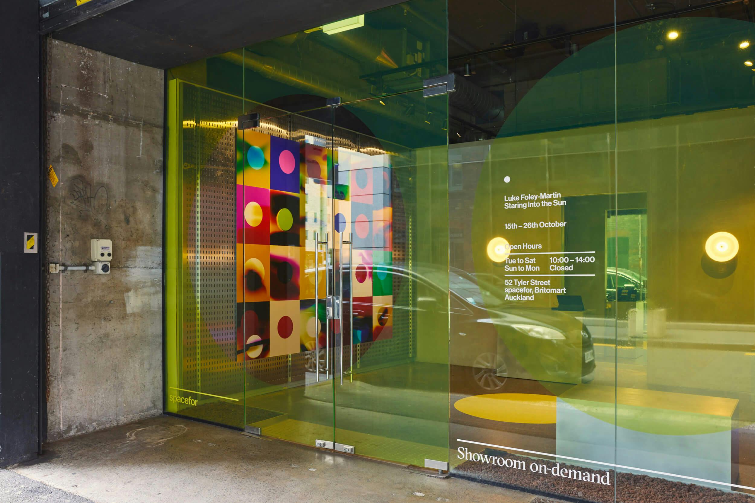 A glass-fronted showroom with green-tinted windows displays colorful polka dot artwork inside. White text on the glass lists exhibition details. A parked car and concrete wall are visible outside.