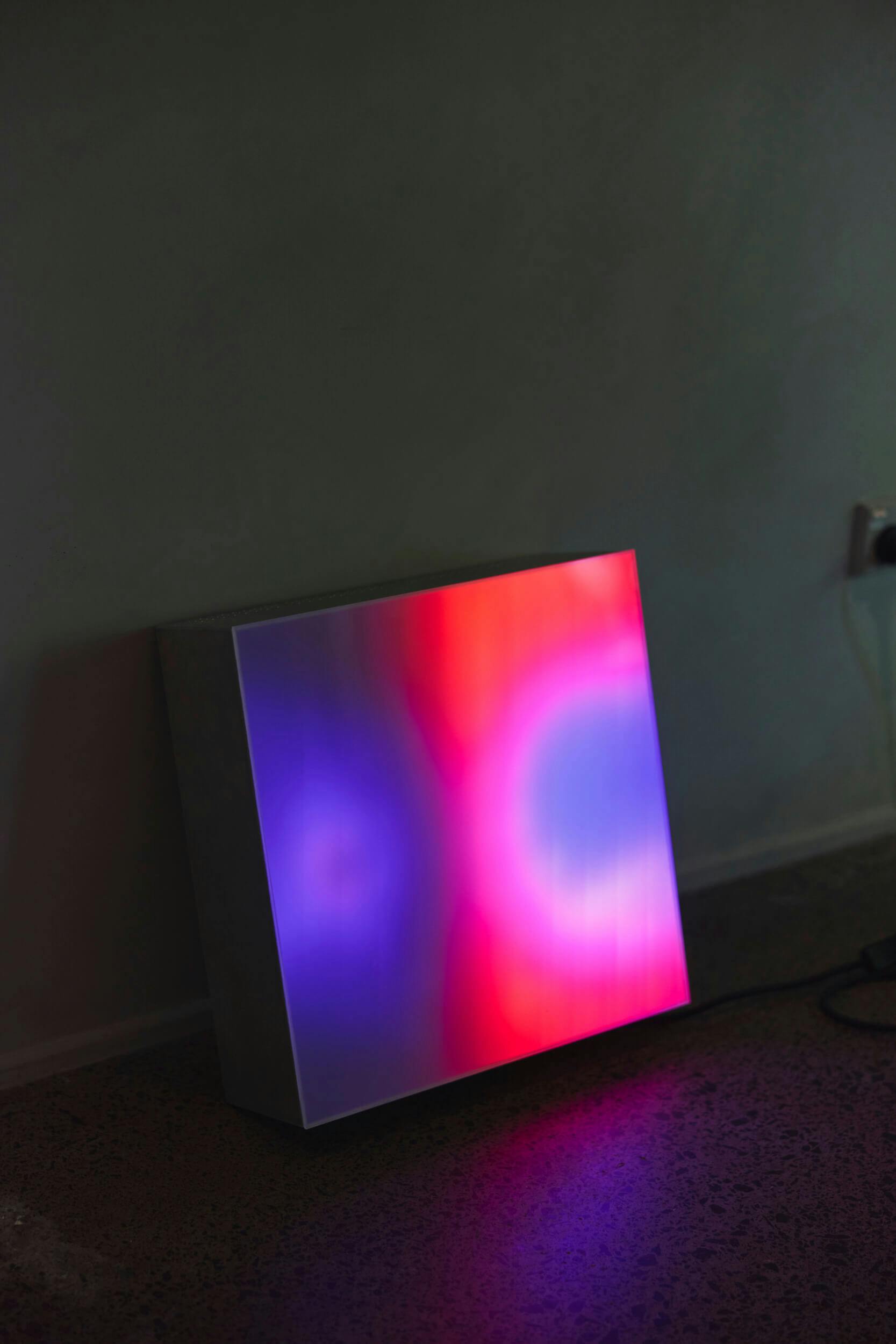 A square lightbox resting on the floor emits a gradient of colorful lights, including purple, pink, blue, and red, casting a glow against a nearby wall and the floor. An electrical outlet and cord are visible in the background.