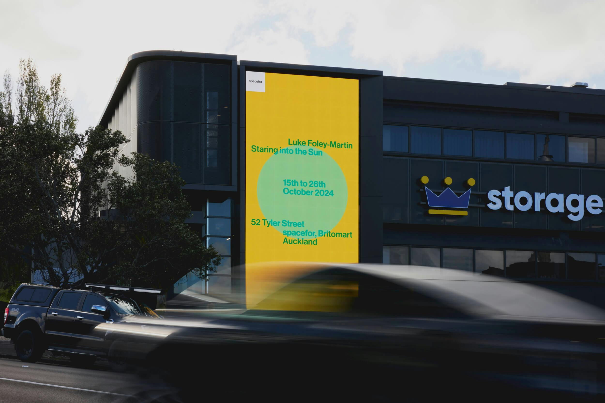 A large digital billboard on the side of a dark building displays a yellow advertisement for an art exhibition by Luke Foley-Martin titled “Staring into the Sun,” scheduled for October 2024 in Auckland. Cars pass in the foreground.