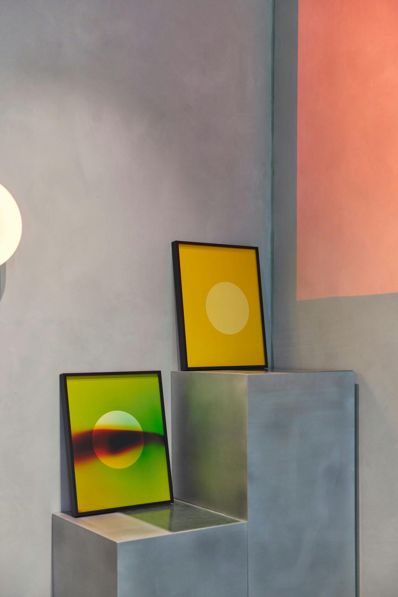 Two framed abstract artworks with circular designs are displayed on metallic pedestals. One artwork has a yellow background with a pale circle; the other has a green and yellow gradient background with a yellow circle.