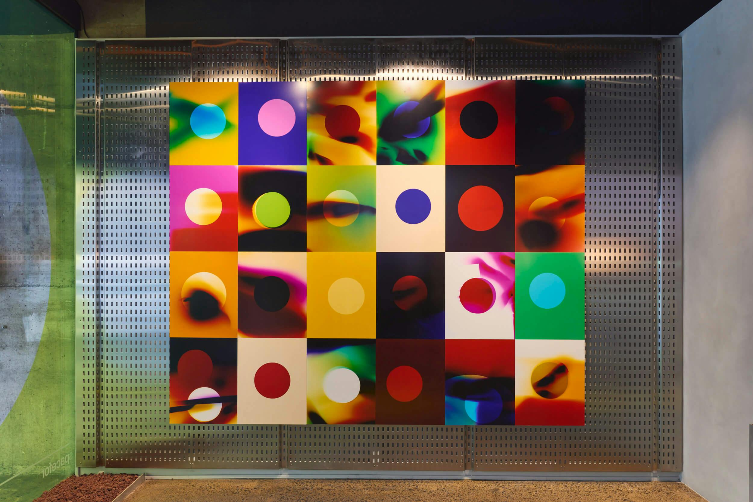 A grid of colorful abstract squares, each with a central circle, is displayed on a metallic perforated wall. The artwork features various vibrant gradients and patterns in each square.