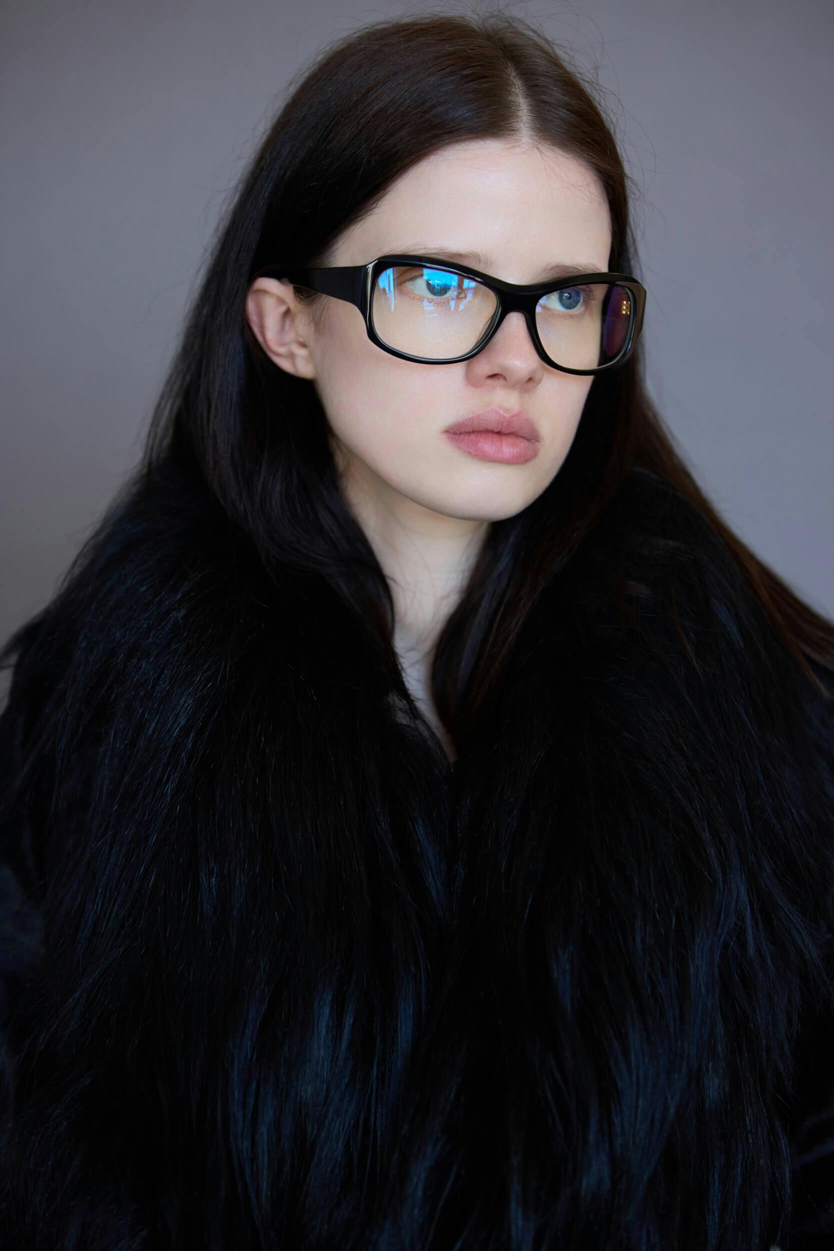 A woman with long dark hair and large black glasses is wearing a black faux fur coat. She is looking slightly to the side against a plain gray background.