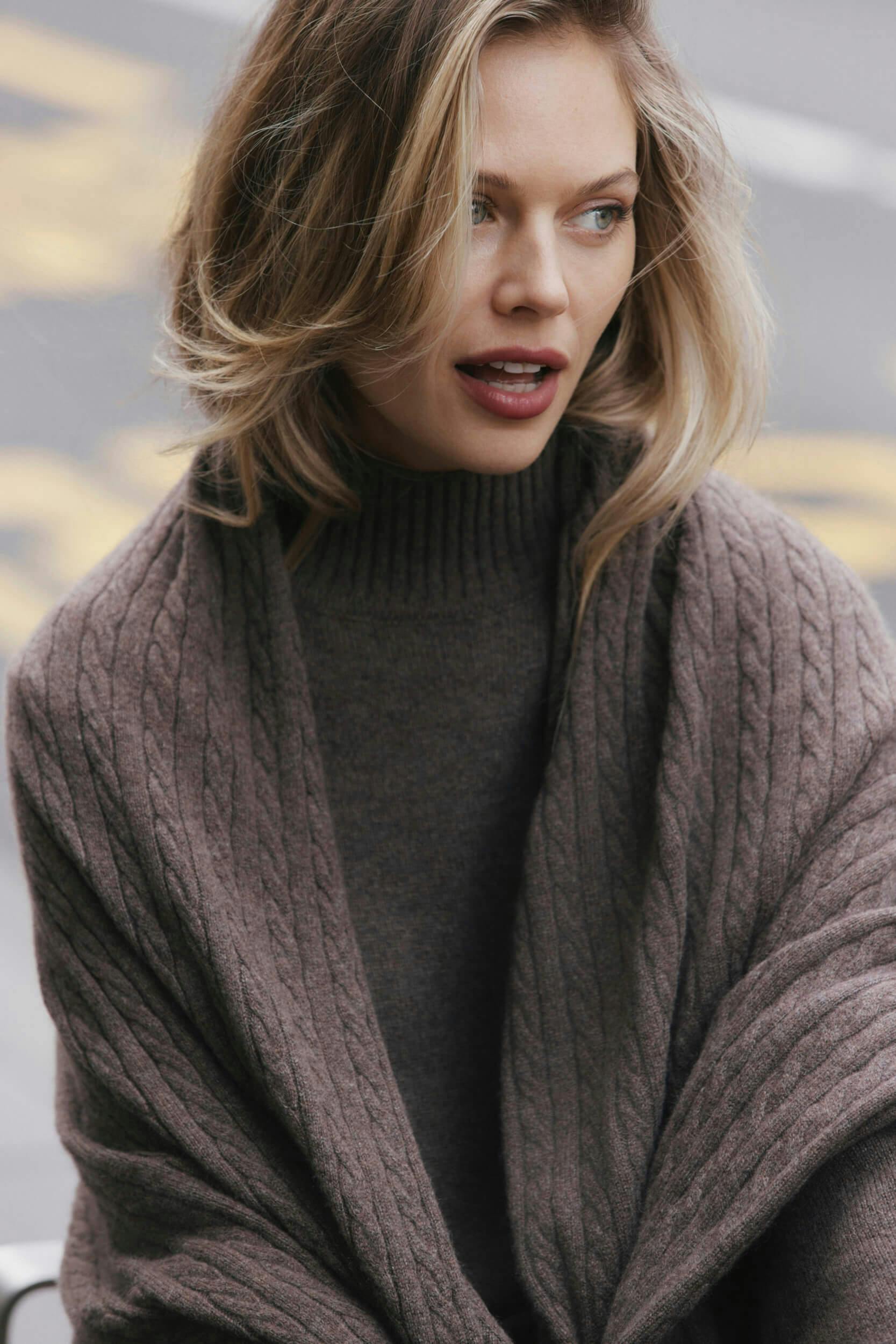 A woman with shoulder-length blonde hair wears a taupe cable-knit shawl over a dark turtleneck sweater. She looks slightly to her left with a neutral expression. The background is blurred and appears to be outdoors.