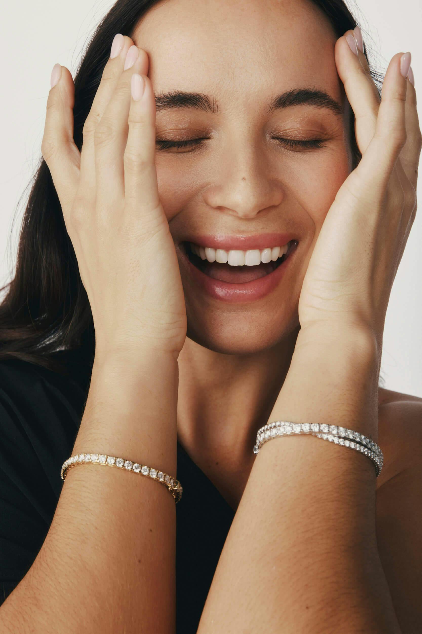 A woman with long dark hair smiles with her eyes closed, holding her face with both hands. She wears a black top and two bracelets, one gold and one silver with stones, on her wrists.