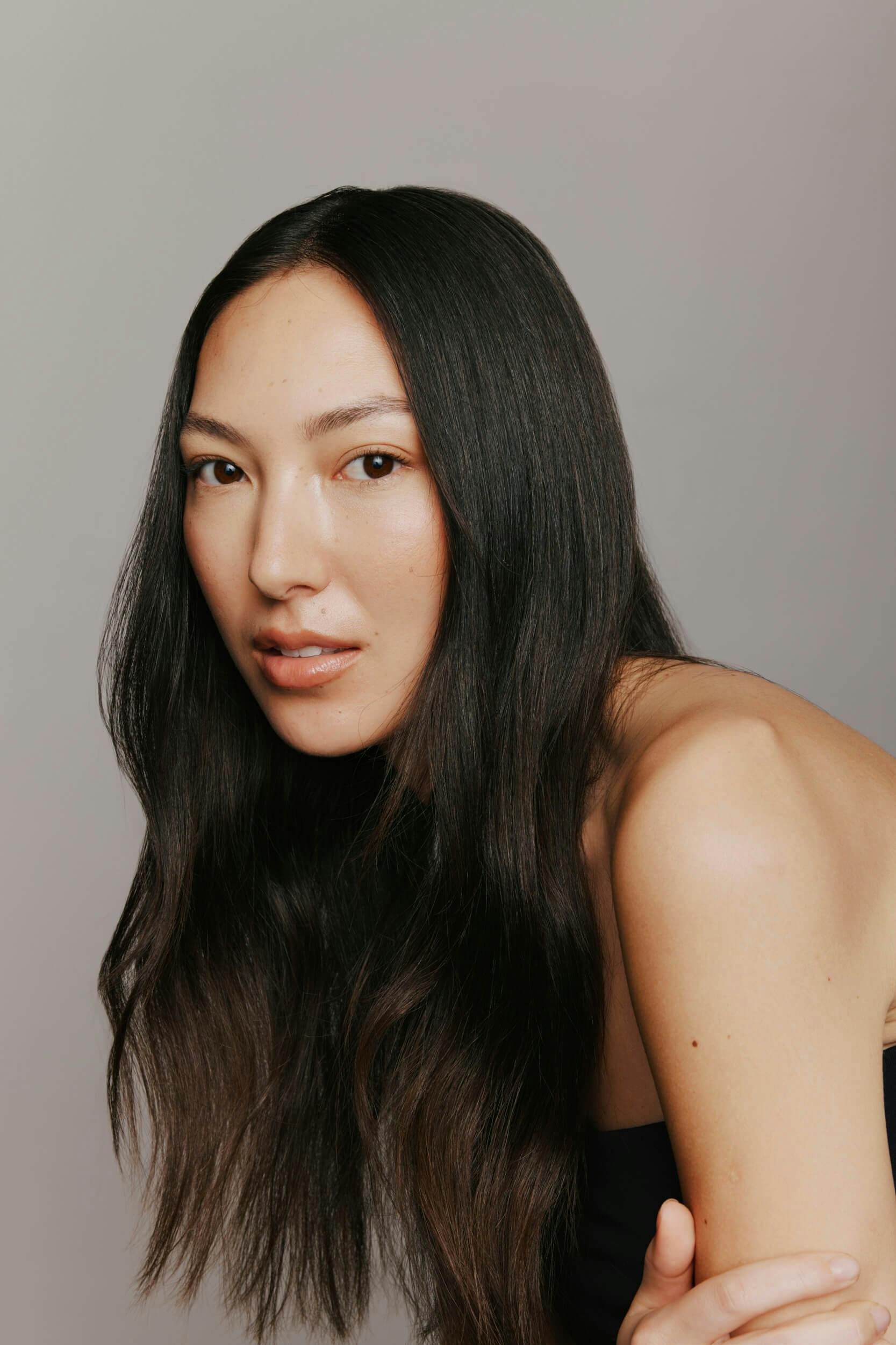 A woman with long, straight dark hair and light skin sits against a plain, neutral background. She has a natural makeup look and is gazing directly at the camera with a neutral expression.