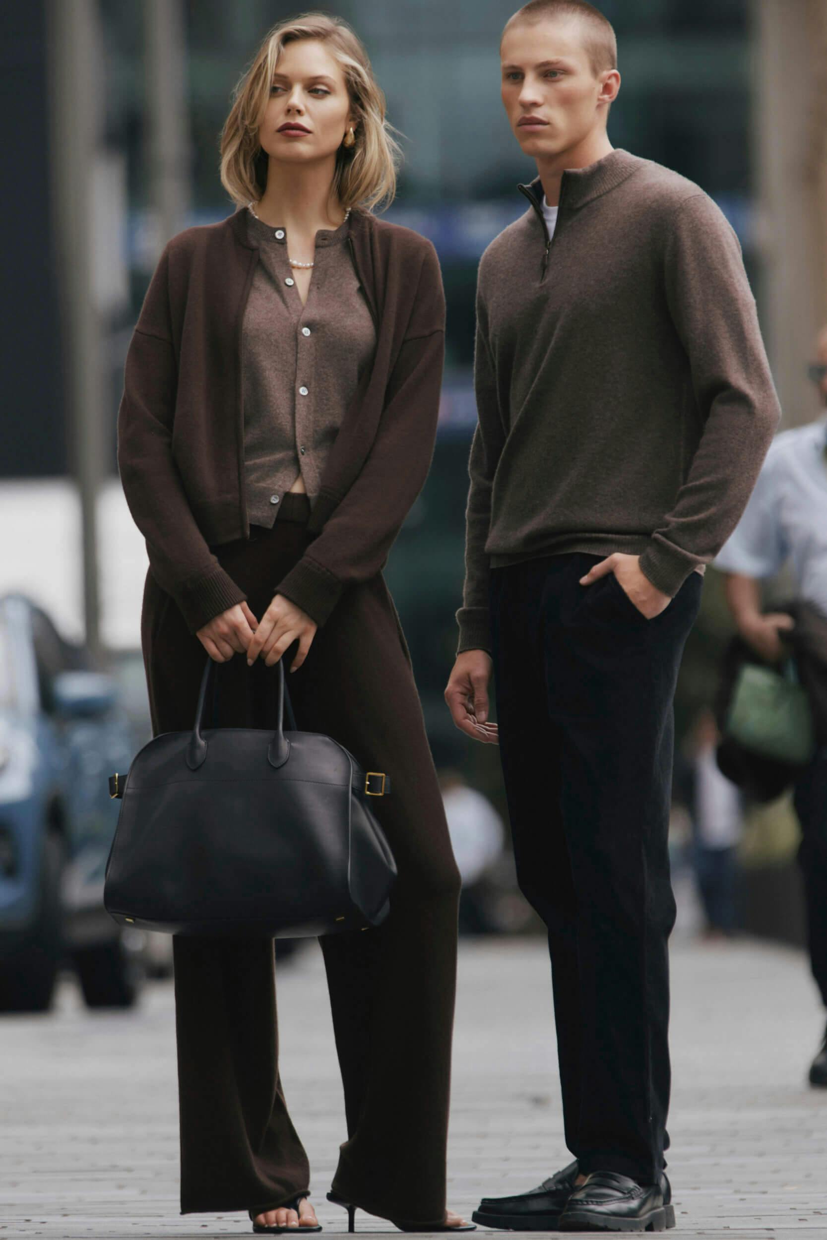 Two models stand side by side outdoors. The woman wears a brown cardigan and pants, holding a large black handbag. The man wears a brown zip sweater and dark trousers. Both look forward with neutral expressions.