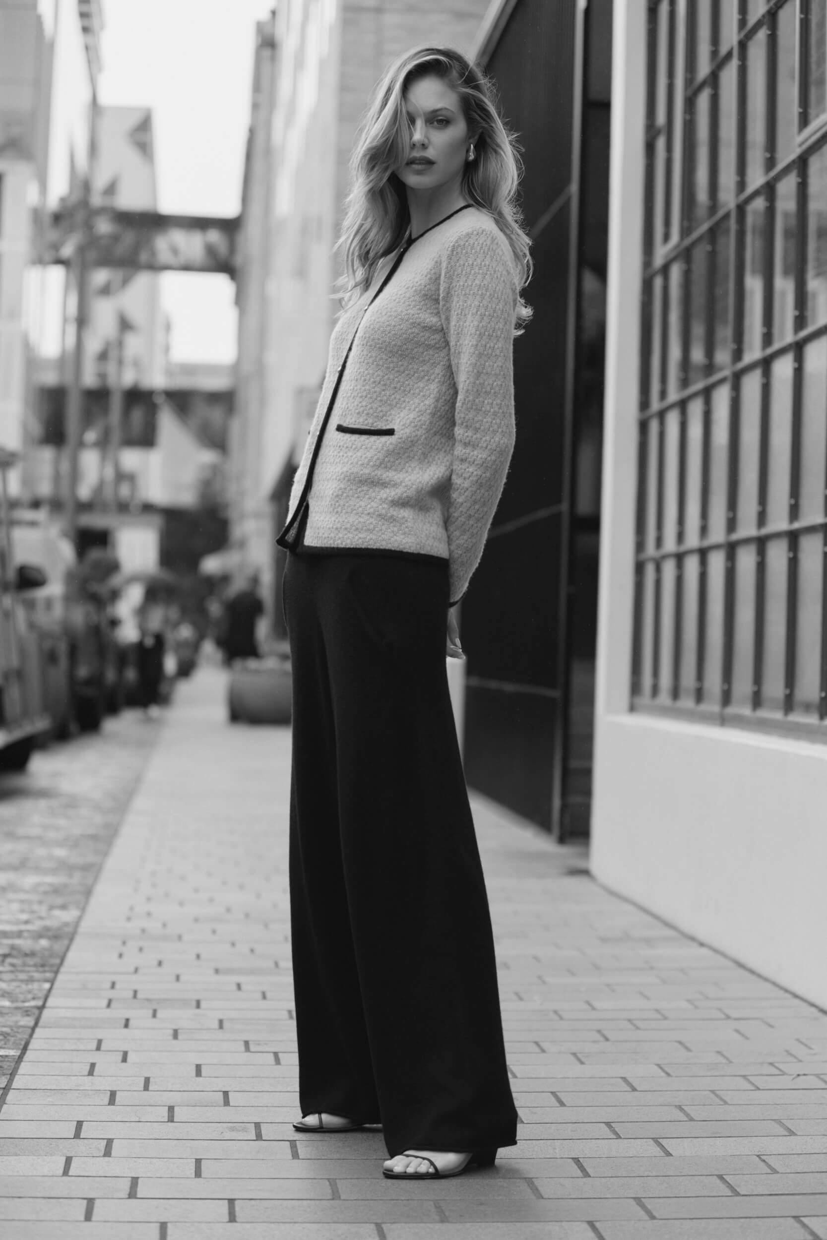 A woman stands on a city sidewalk wearing a light-colored textured jacket, wide-leg dark trousers, and open-toe shoes. She looks toward the camera with buildings and parked cars in the background. The image is in black and white.