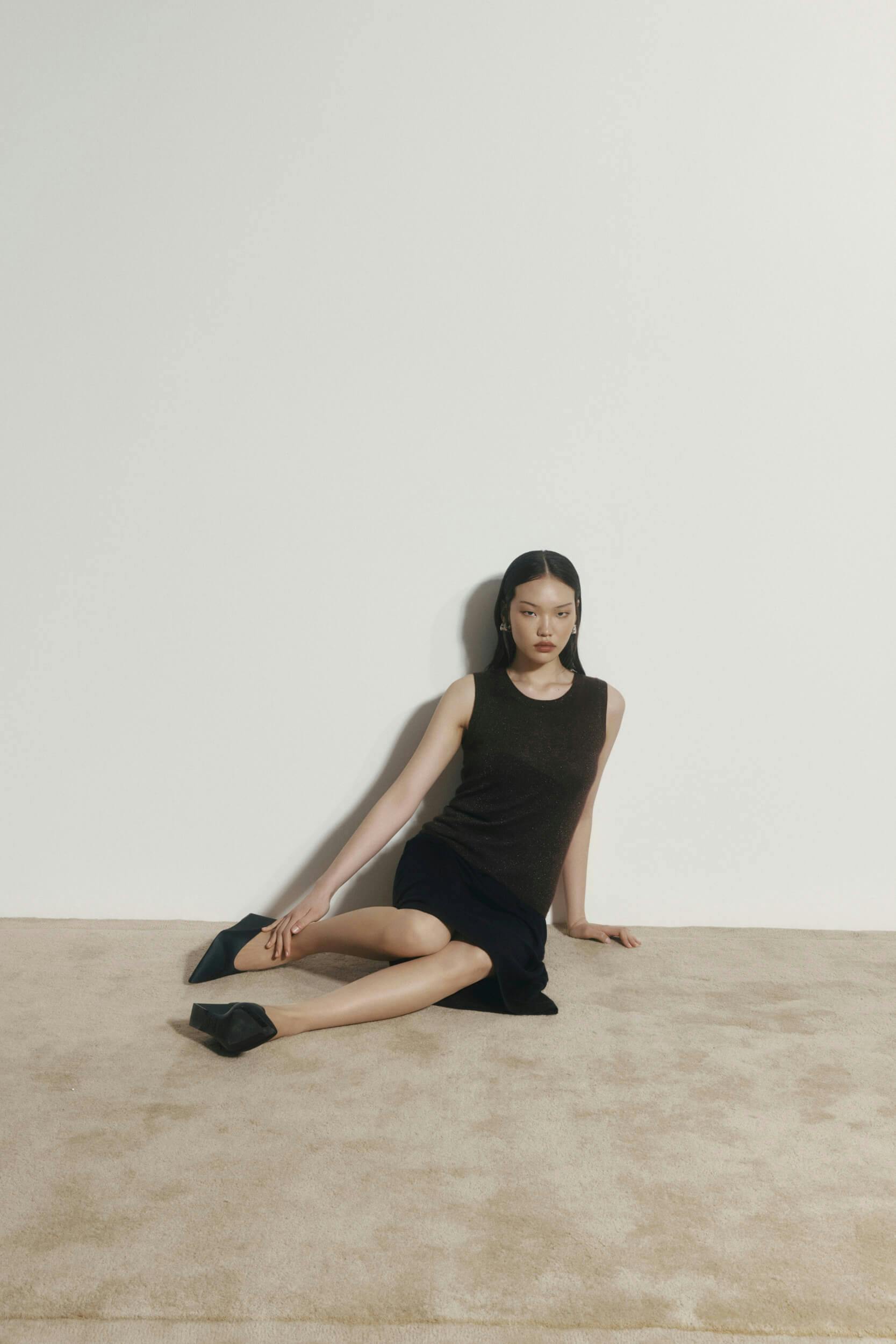 A woman with straight black hair sits on a beige carpet against a white wall, wearing a sleeveless dark top, black skirt, and black shoes, with one leg bent and one hand resting on the floor.