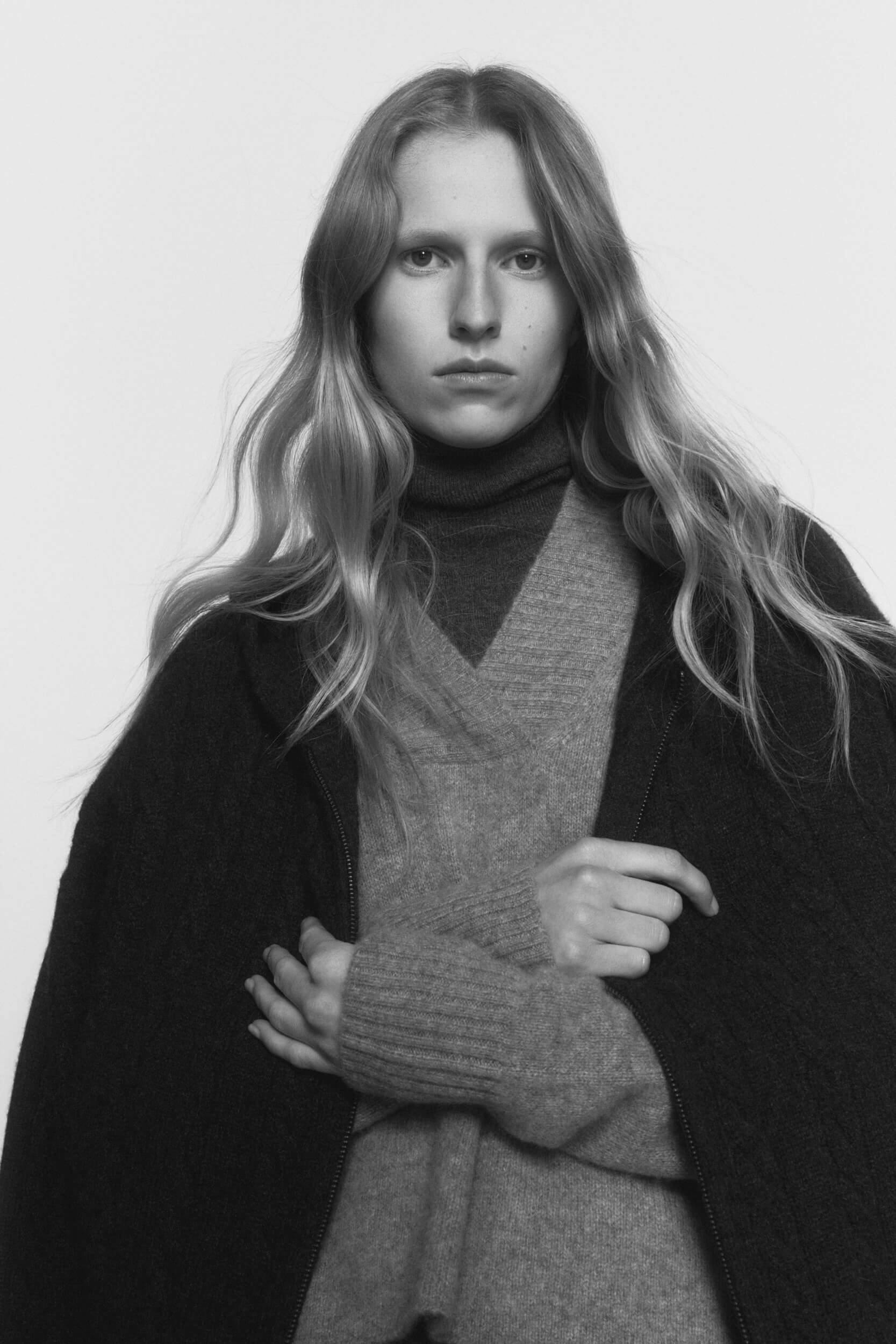 A person with long, wavy hair poses against a plain background, wearing a dark textured coat over a high-neck sweater and a v-neck sweater, looking directly at the camera with a neutral expression.