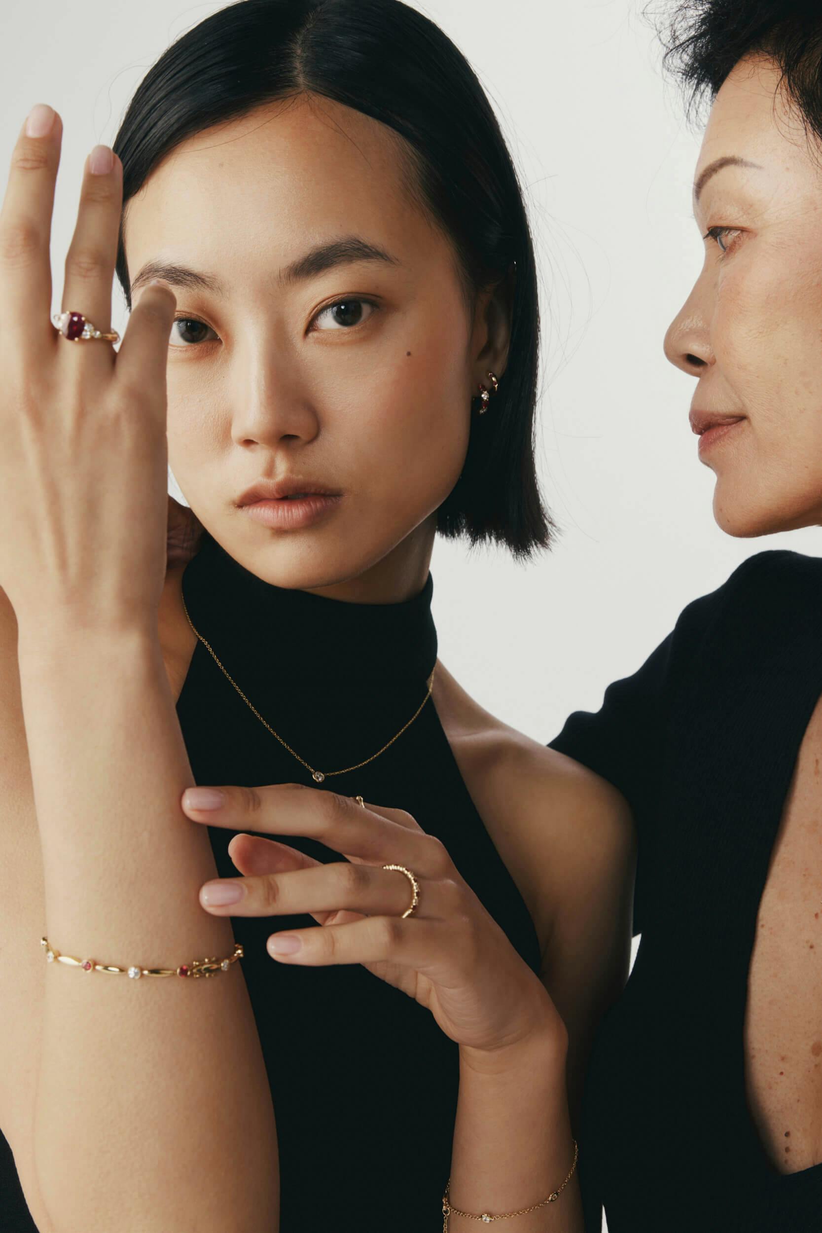Two women standing close together, both wearing black tops and gold jewelry including rings, a bracelet, and a necklace. The woman in front looks at the camera while the other faces her in profile against a plain background.