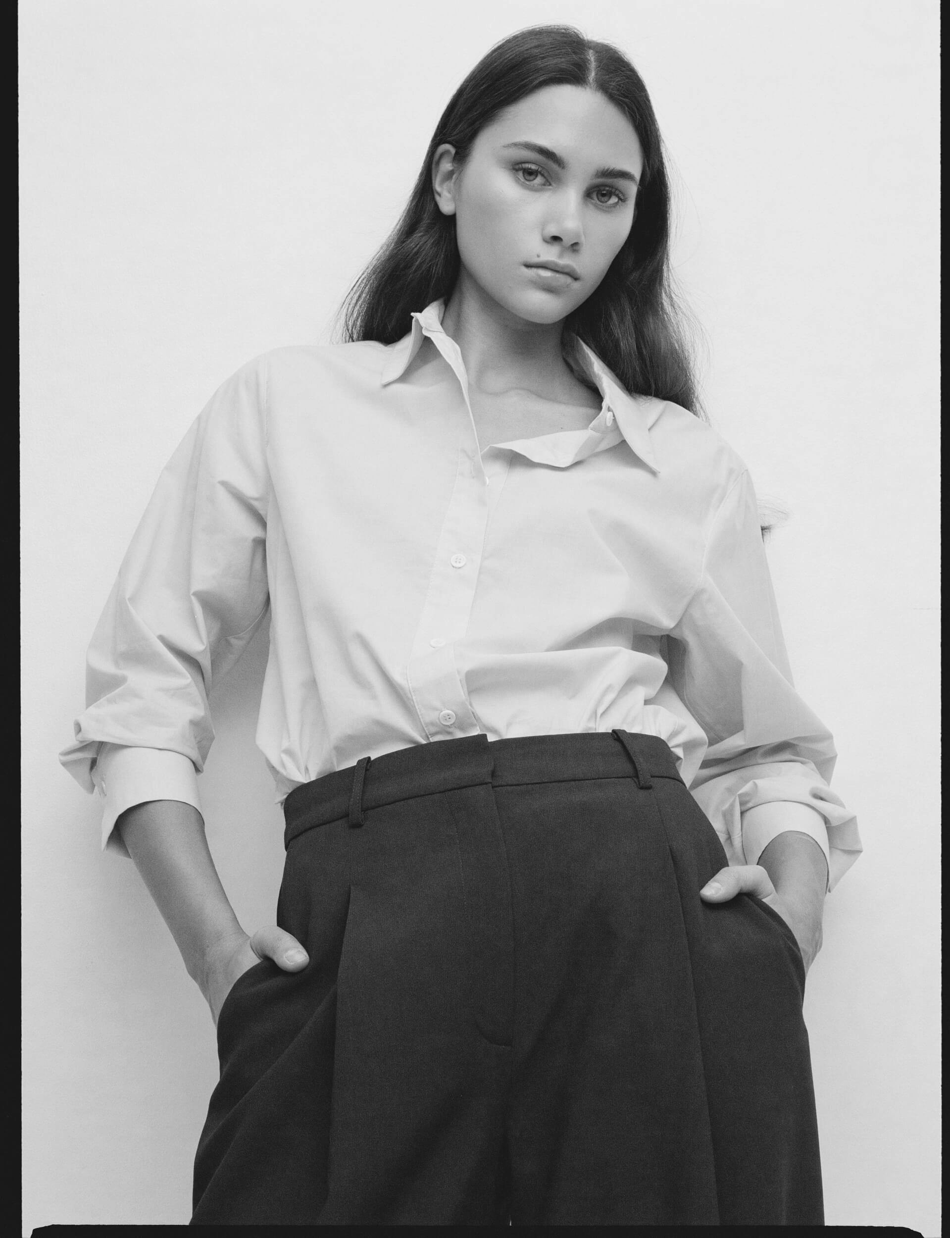 A person with long, straight hair is wearing a light button-up shirt tucked into high-waisted, dark trousers. They stand against a plain background with their hands in their pockets, looking at the camera.