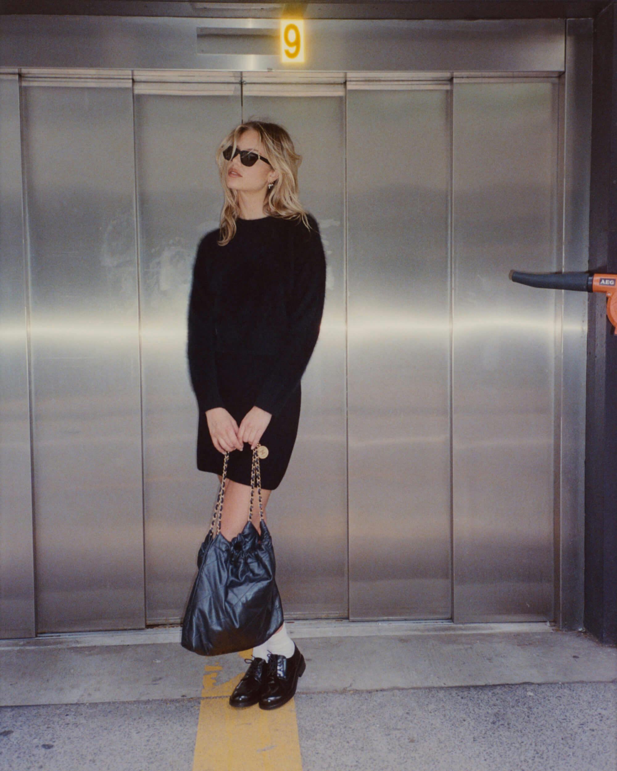 A person wearing sunglasses, a black sweater, a black skirt, and black shoes with white socks stands in front of closed elevator doors, holding a large black bag with a chain strap. A yellow number 9 is above the doors.