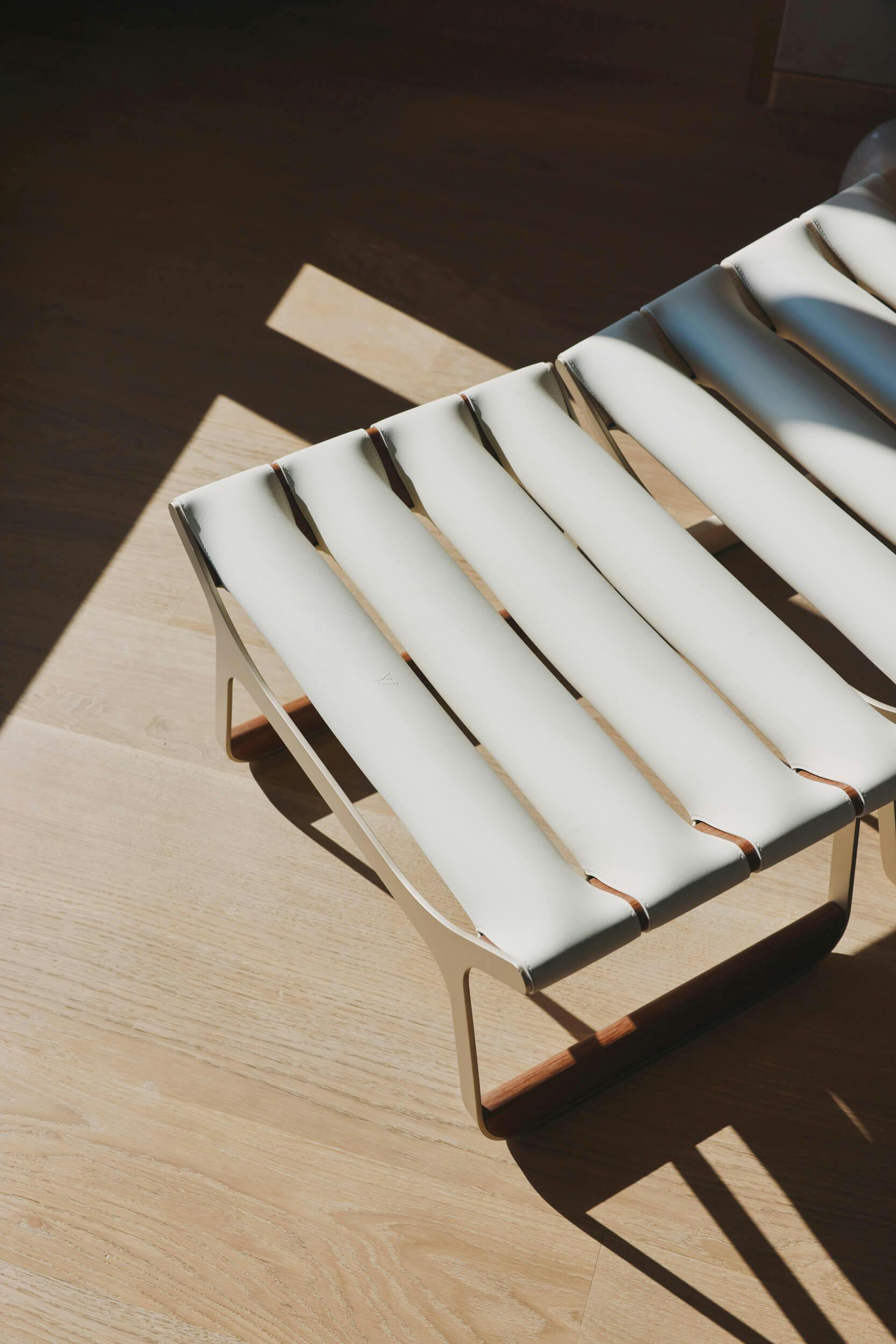 A modern bench with white, padded slats and metal legs stands on a light wooden floor, illuminated by natural sunlight coming from the left.